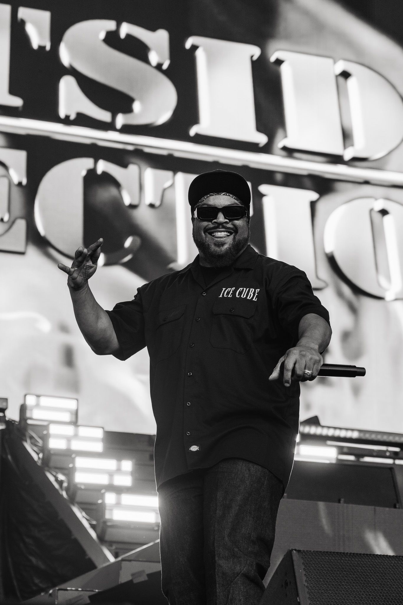 Black and white photo of Ice Cube performing on stage, smiling, holding a microphone, wearing sunglasses and a cap.