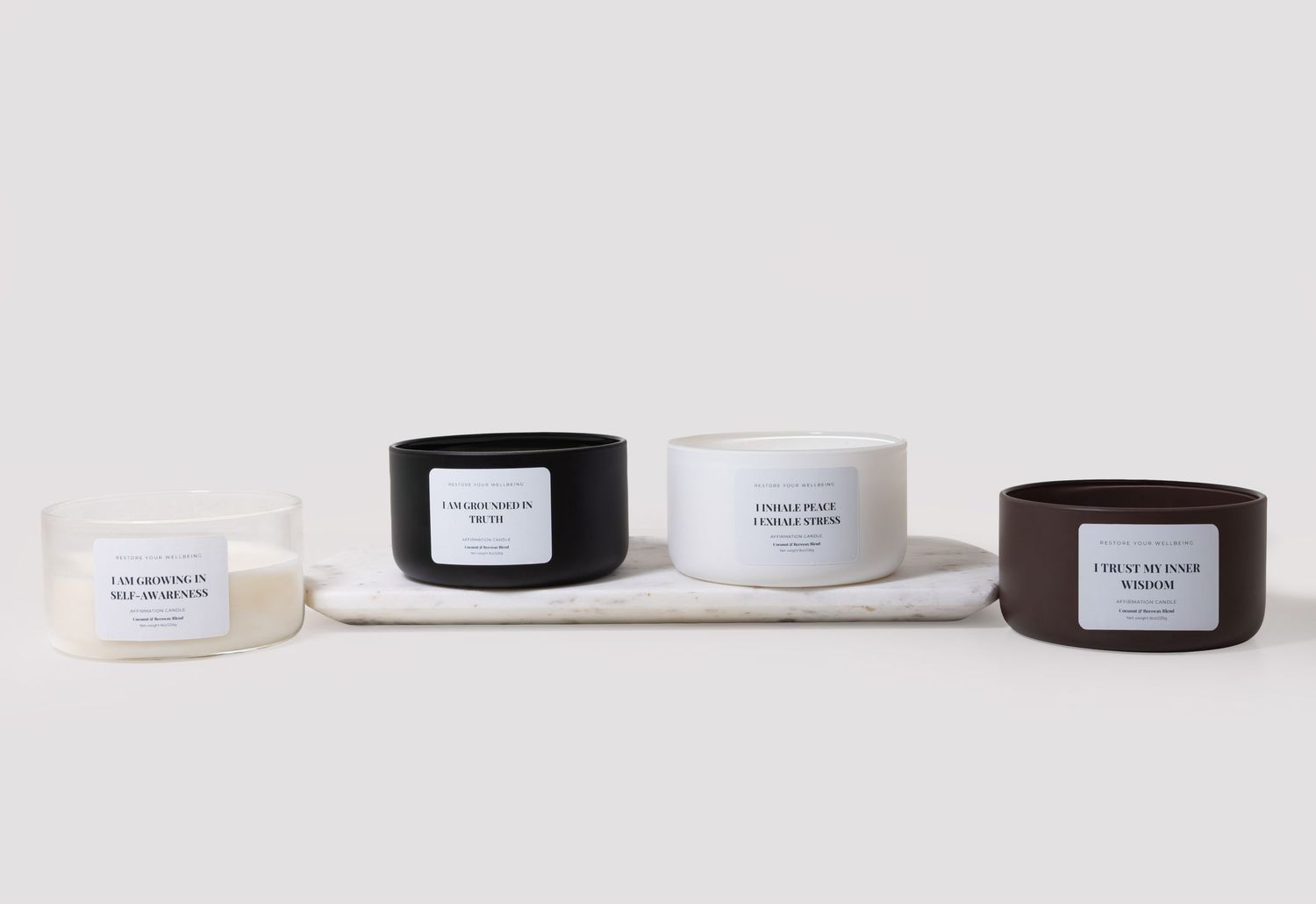 Four scented candles in cream, black, white, and brown containers with label text on a marble tray.