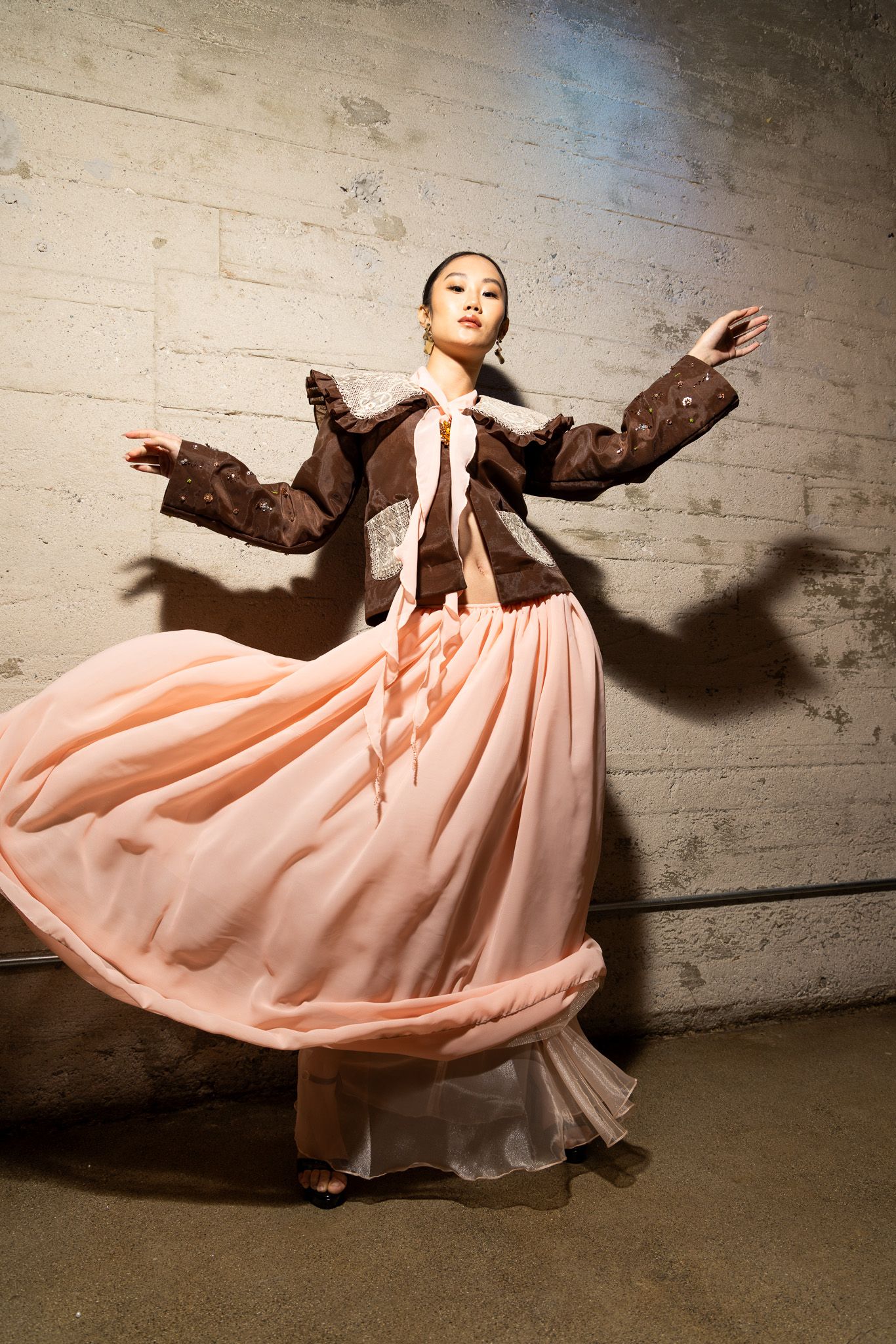 A dancer in a flowing pink skirt and brown jacket twirls gracefully against a concrete wall in a dynamic motion sequence.