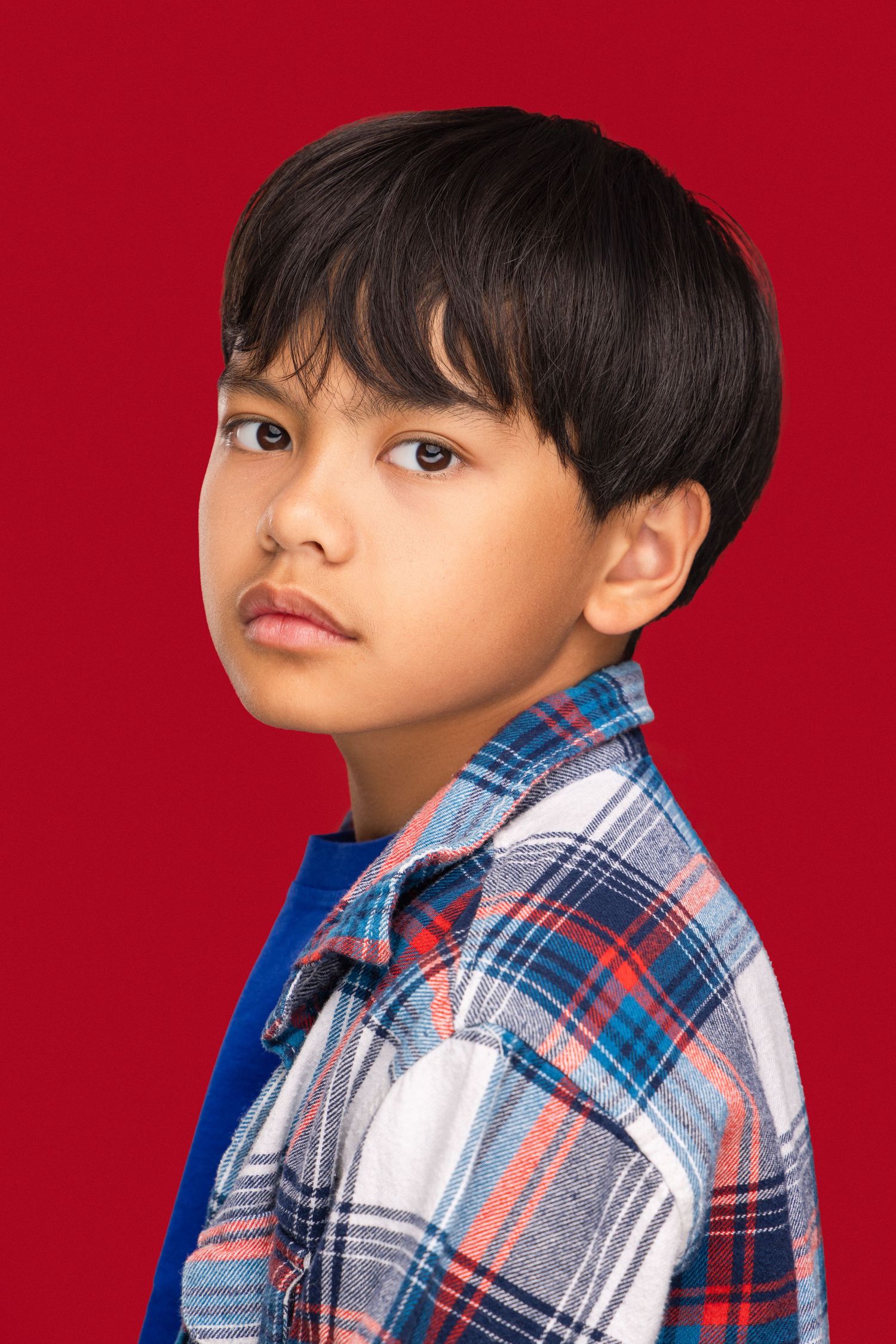 Child wearing plaid button-up shirt poses against red background in studio portrait series.