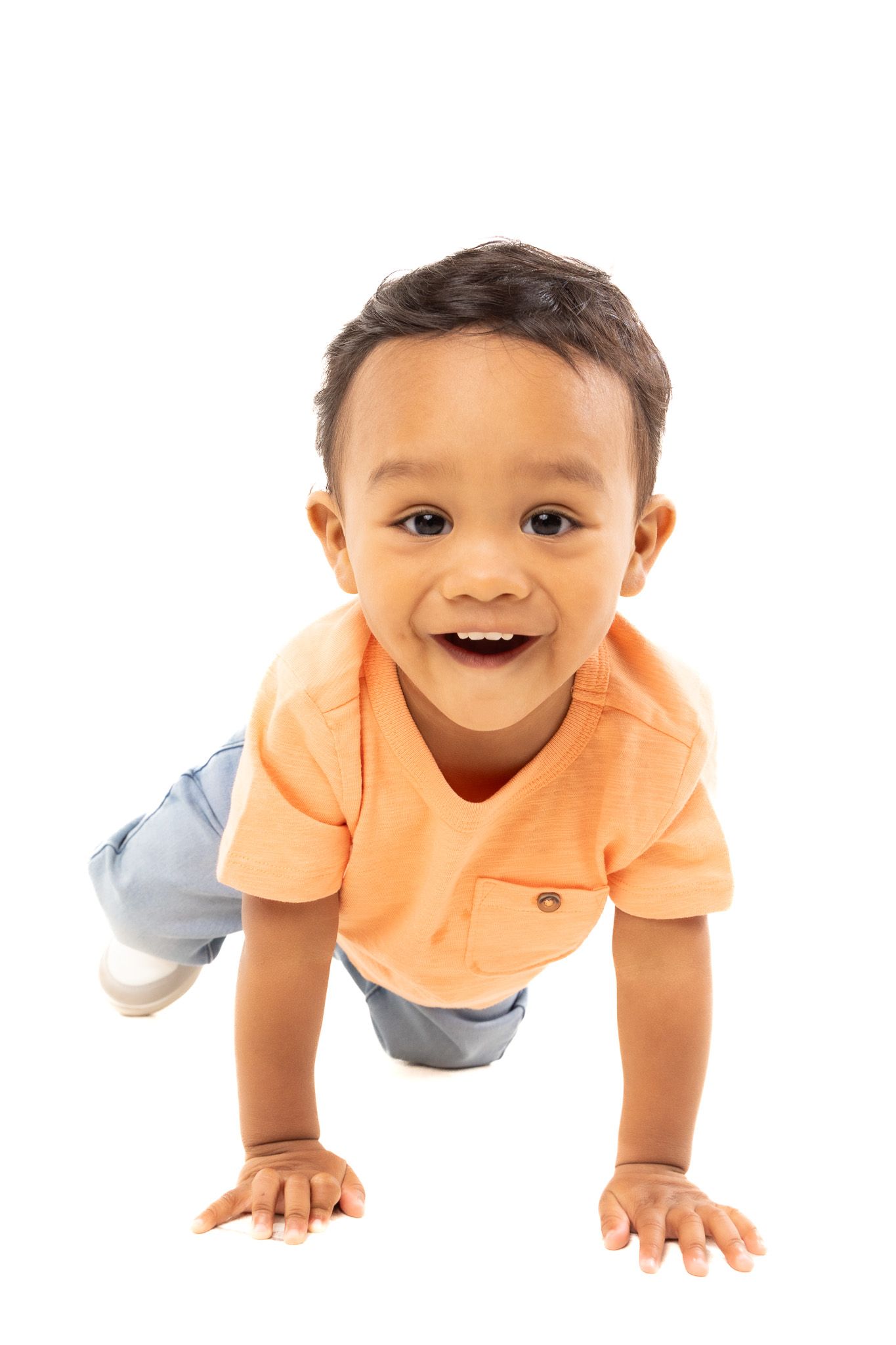 Smiling baby in orange shirt and gray pants crawling on white background.