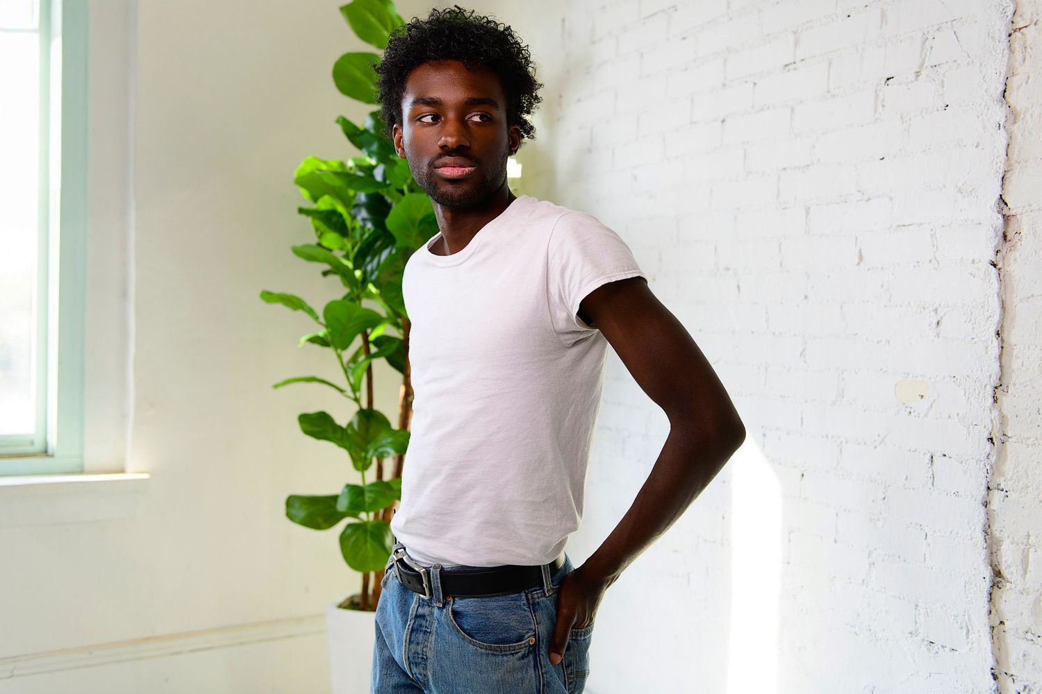 Figure in a white t-shirt and jeans poses casually against a bright wall with green plants nearby.