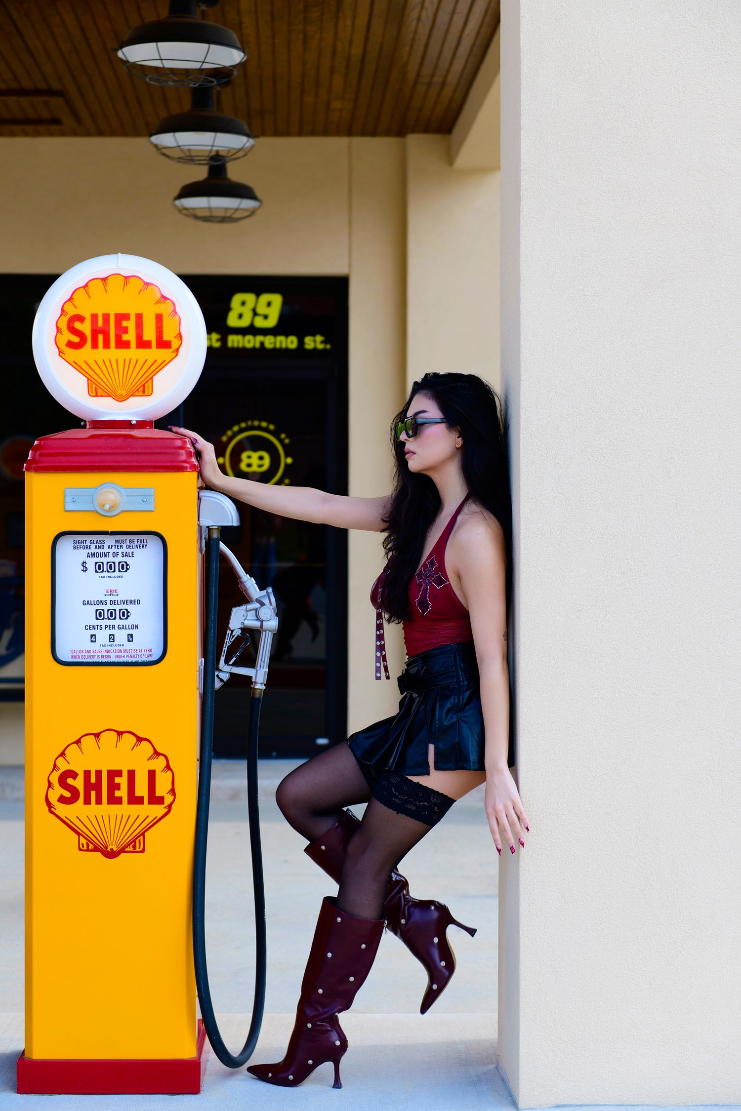 Stylized pinup photoshoot with vintage yellow Shell gas pump featuring high heels and fishnet stockings.