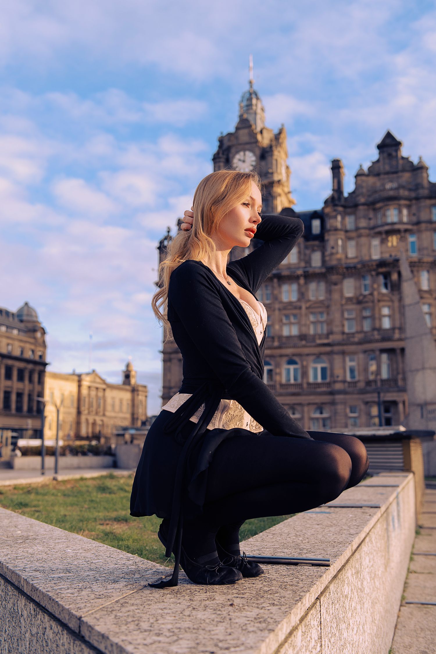 Person in black outfit sits on stone wall near historic Gothic architecture building under blue cloudy sky.