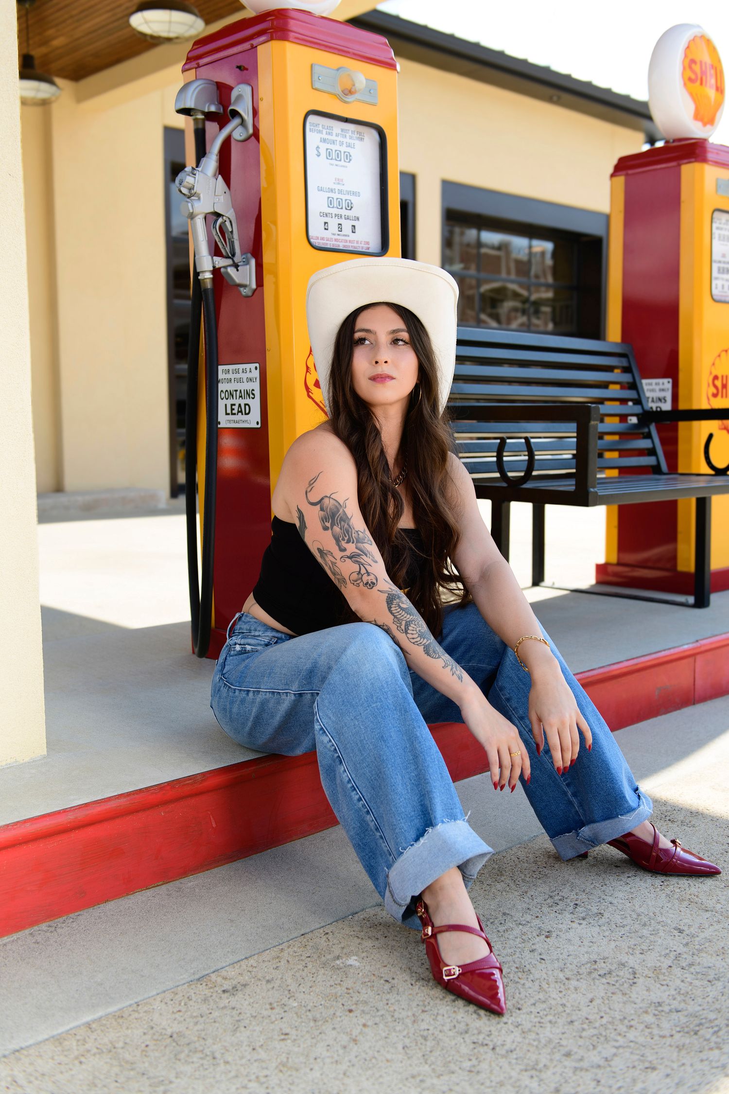 A person in jeans and burgundy shoes sits casually on the ground against a retro-style gas station pillar.