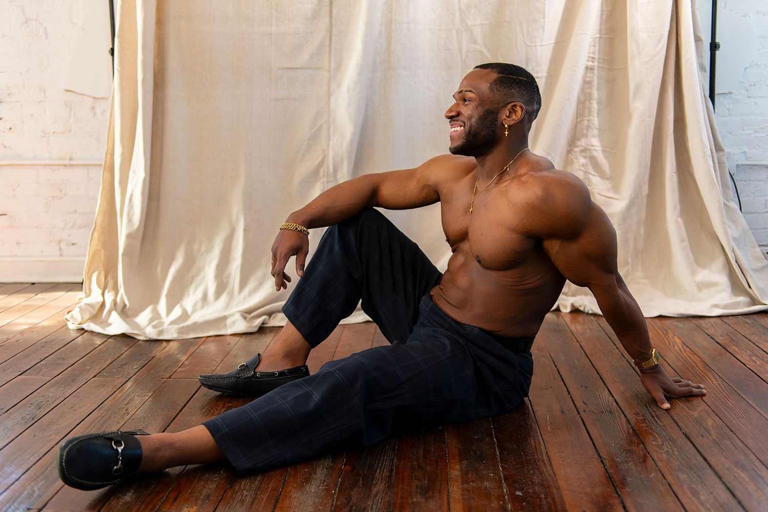 Athletic person in black pants sits on wooden floor against white curtain backdrop during fitness photoshoot.