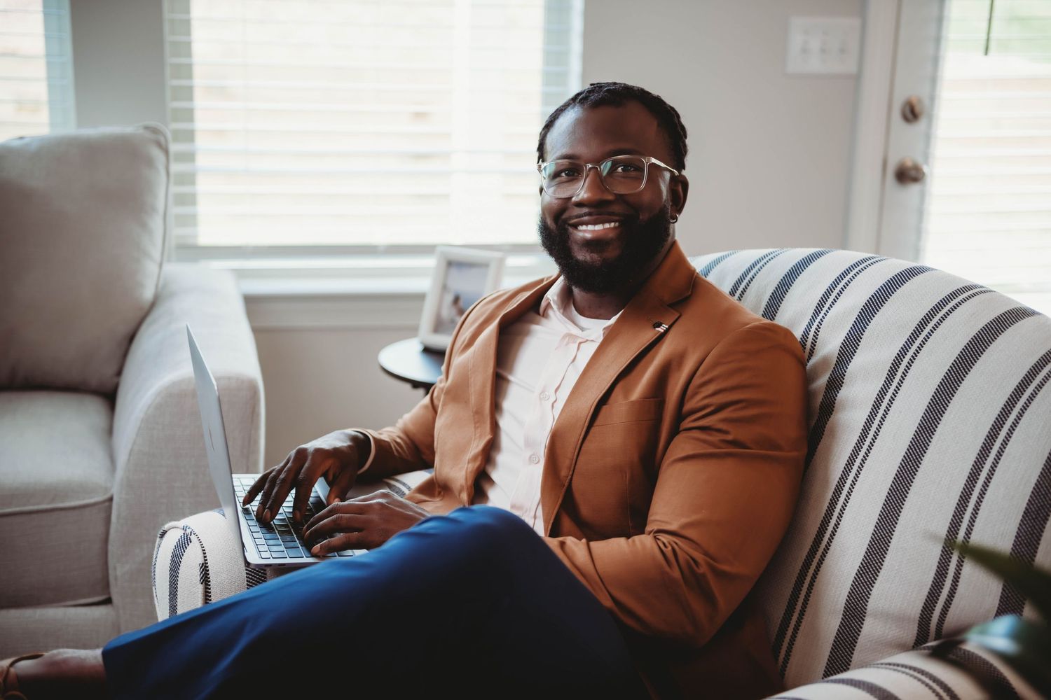 Professional relaxed on a striped couch wearing a brown blazer in natural window light.
