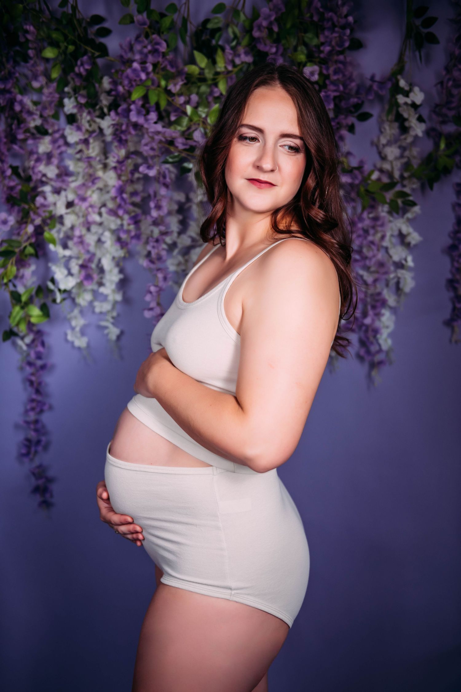 Maternity portrait against purple wisteria backdrop in soft lighting with white lingerie.