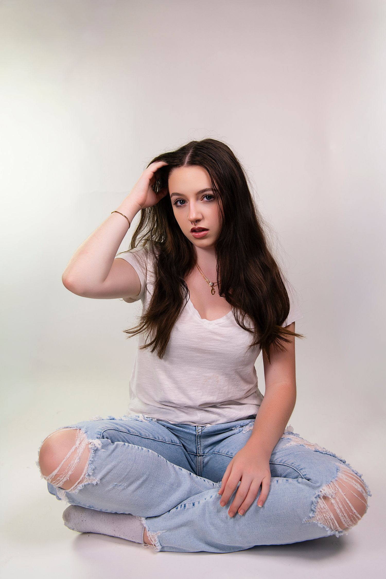 A person in ripped light blue jeans and white tank top poses casually on a light studio backdrop.