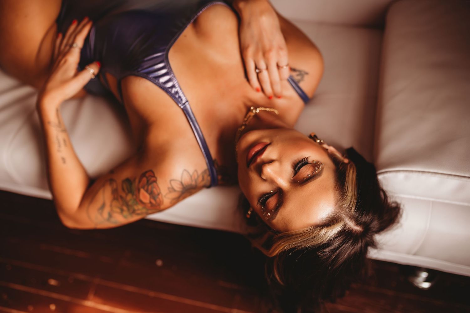 Woman in blue lingerie lying on a sofa, showcasing tattoos on her arm, in a boudoir photography style.