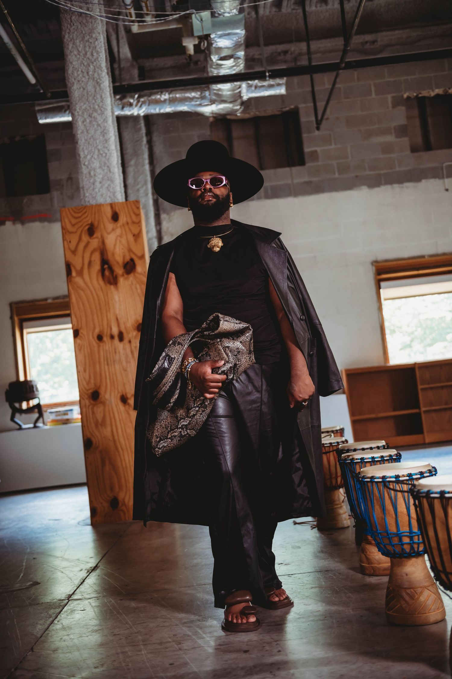 A figure in a dramatic black cape, wide-brimmed hat and red sunglasses stands in a moody industrial interior space.