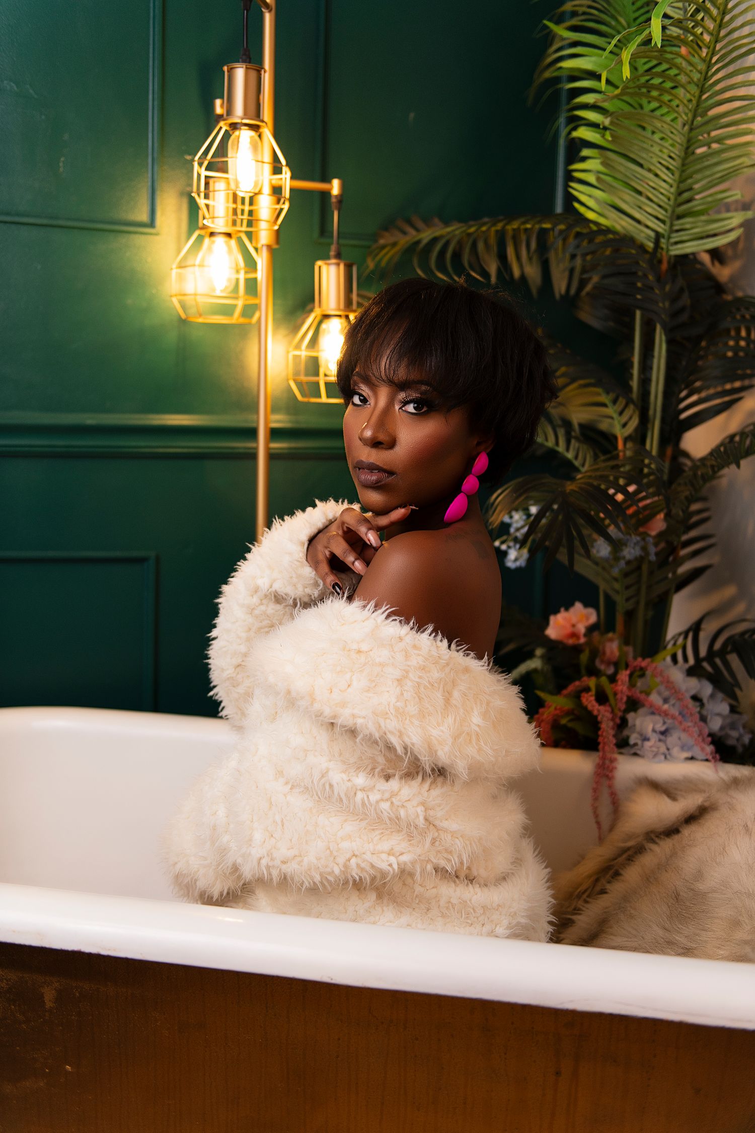 A person in a white fuzzy robe relaxes in a luxurious bathtub with tropical plants and warm lighting in the background.