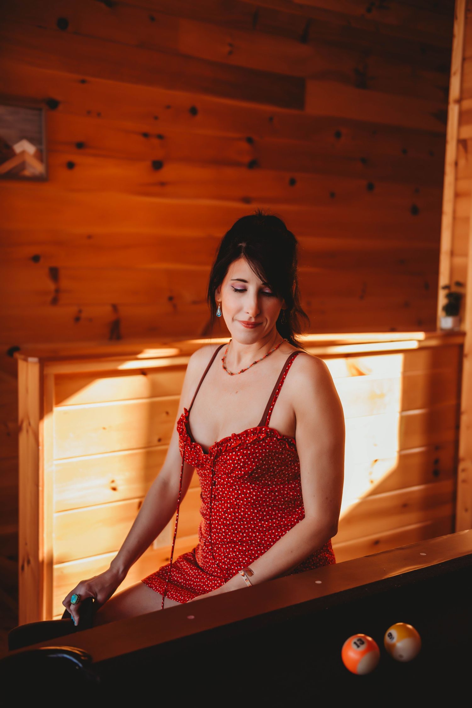 Playing pool in a rustic cabin wearing a red polka dot sundress in warm lighting.