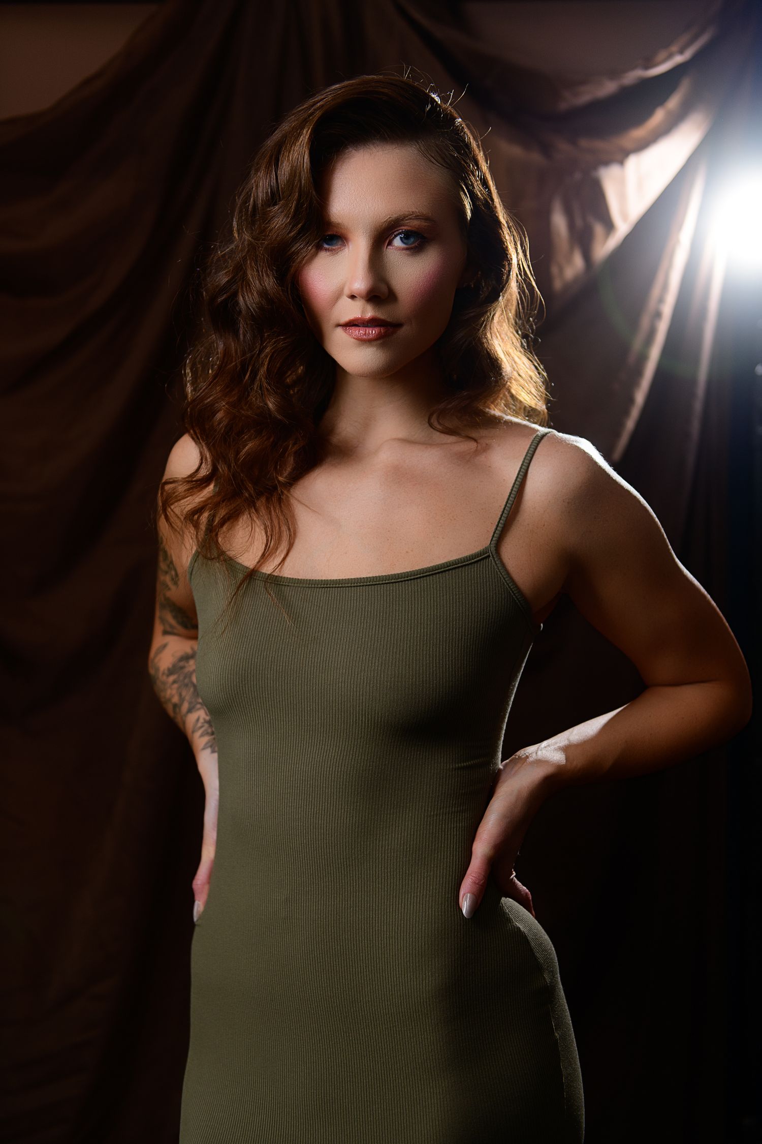 A figure in an olive green sleeveless top poses dramatically against a dark background with dramatic rim lighting.