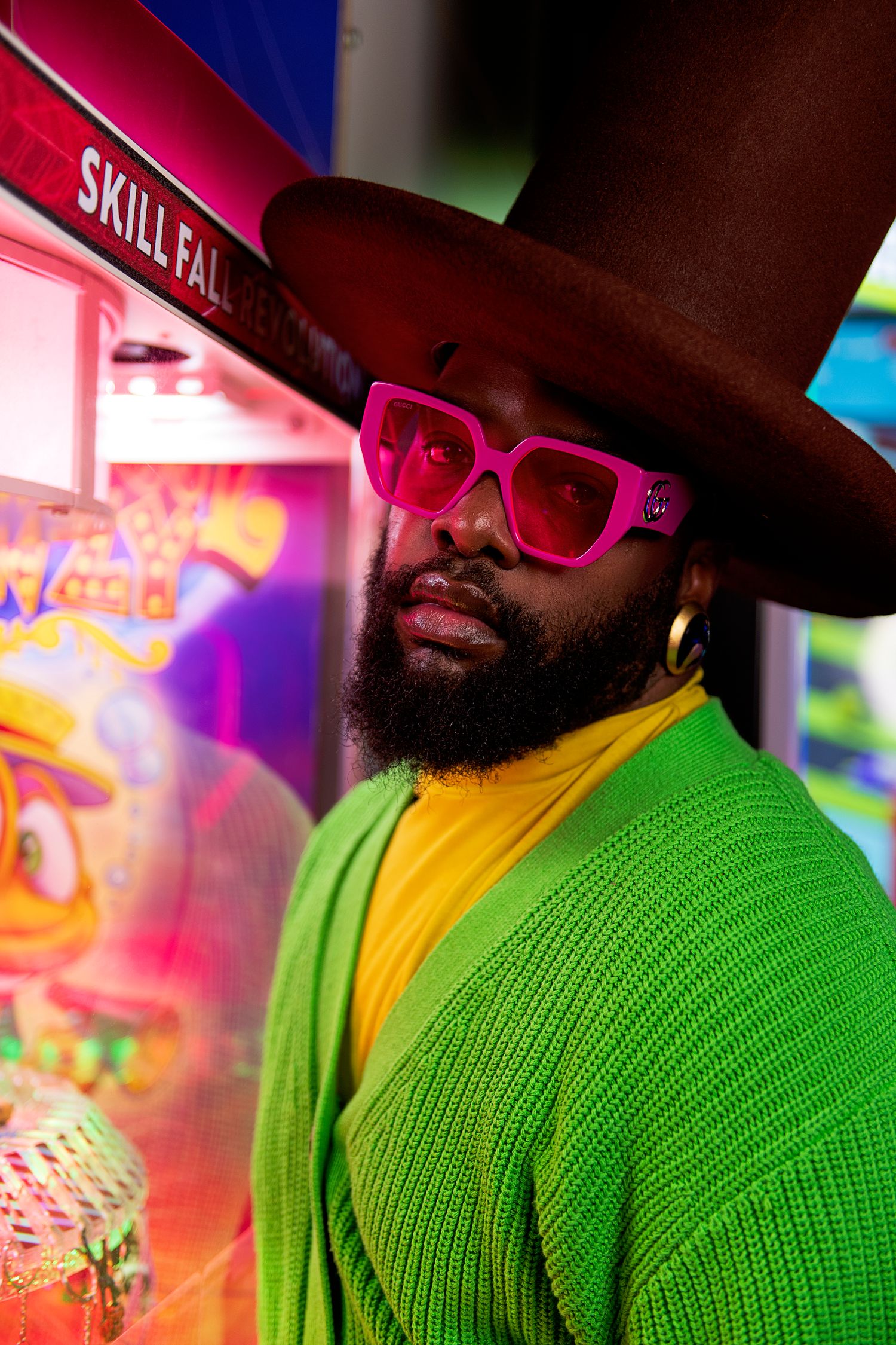 Stylish person in brown hat and pink sunglasses wearing bright green cardigan against colorful arcade background.