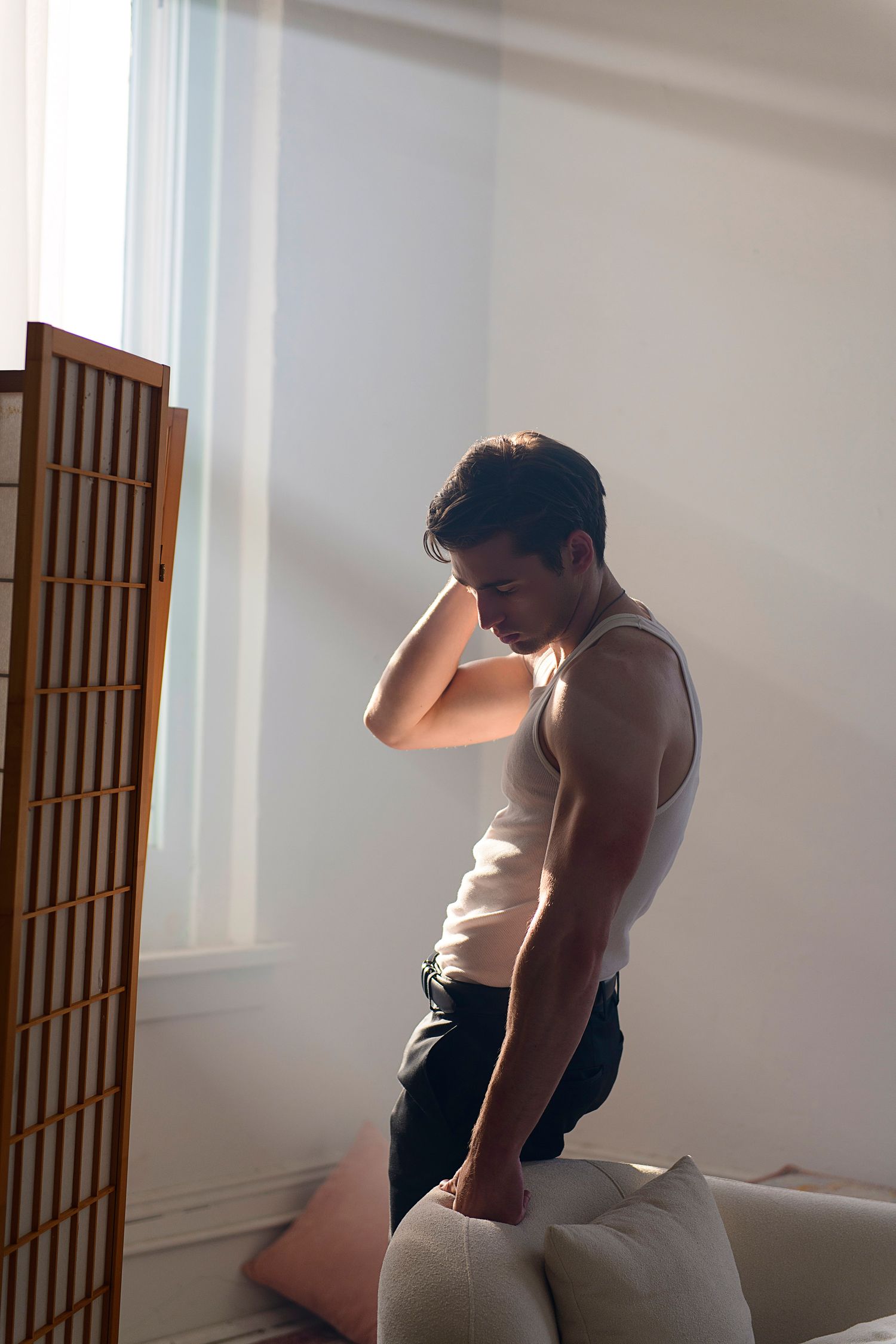 Person in white tank top and dark bottoms sits contemplatively by a window with dramatic natural lighting.