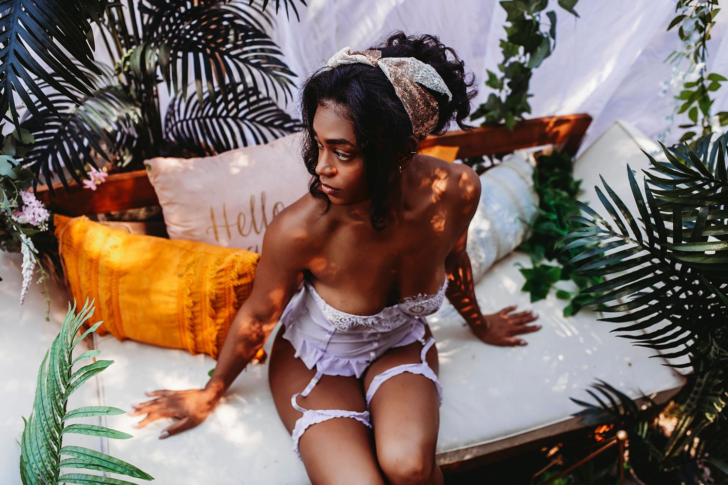 Woman in white lingerie lounges on bed with tropical plants and warm lighting creating sensual boudoir atmosphere.