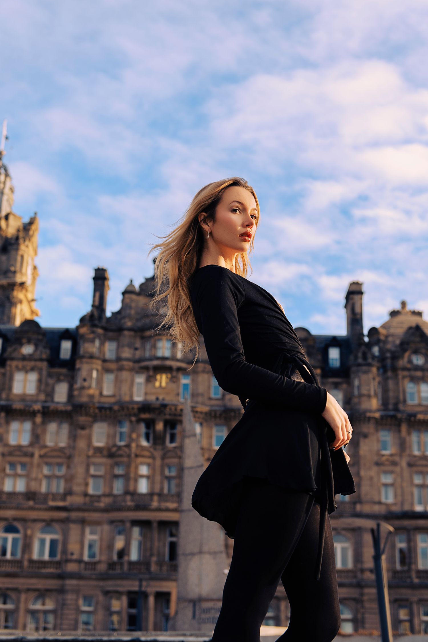 A figure in a black coat poses dramatically against historic Edinburgh architecture with cloudy skies in the background.