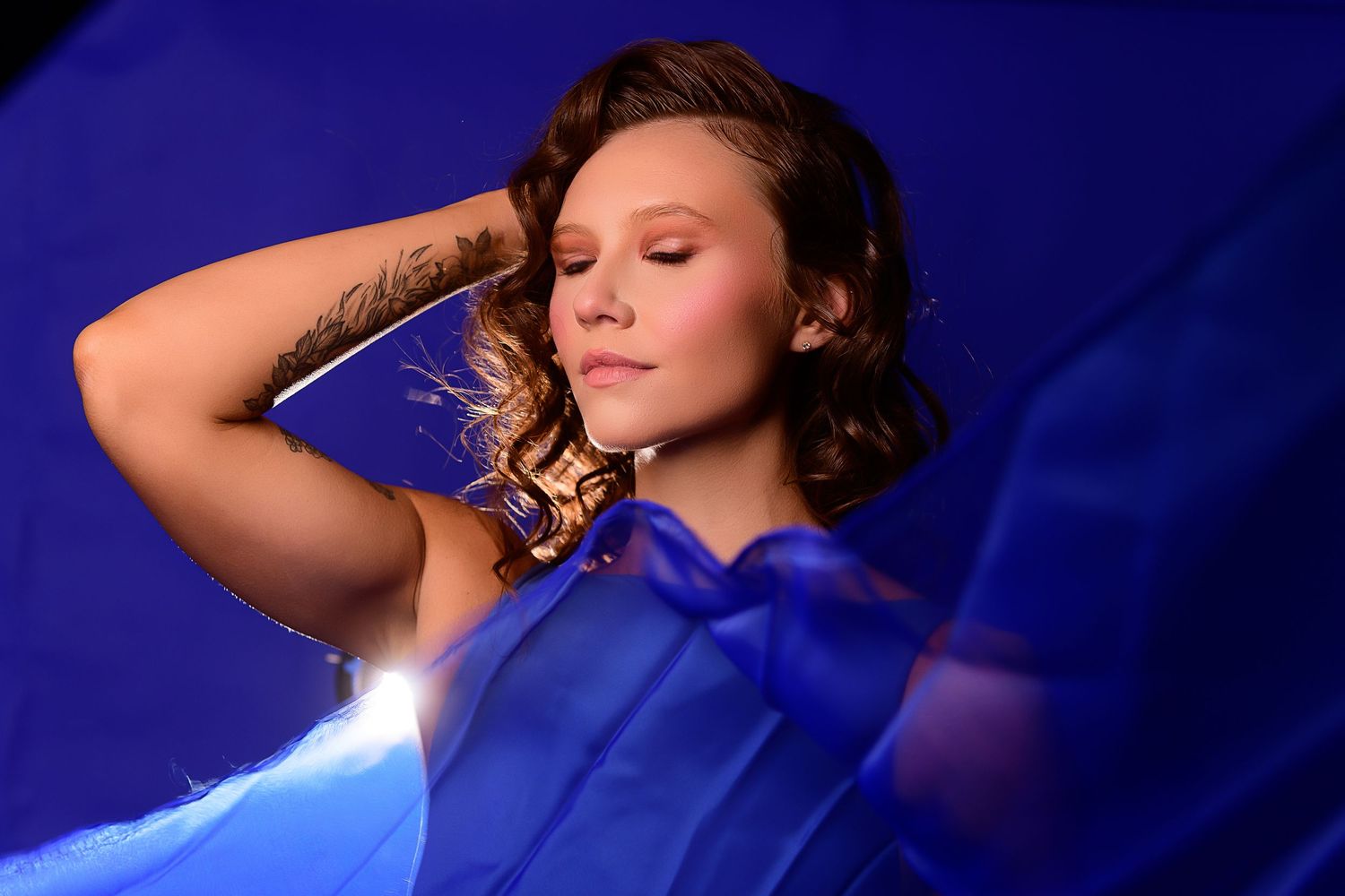 Woman in blue dress poses elegantly against dark background in artistic glamour photoshoot series.