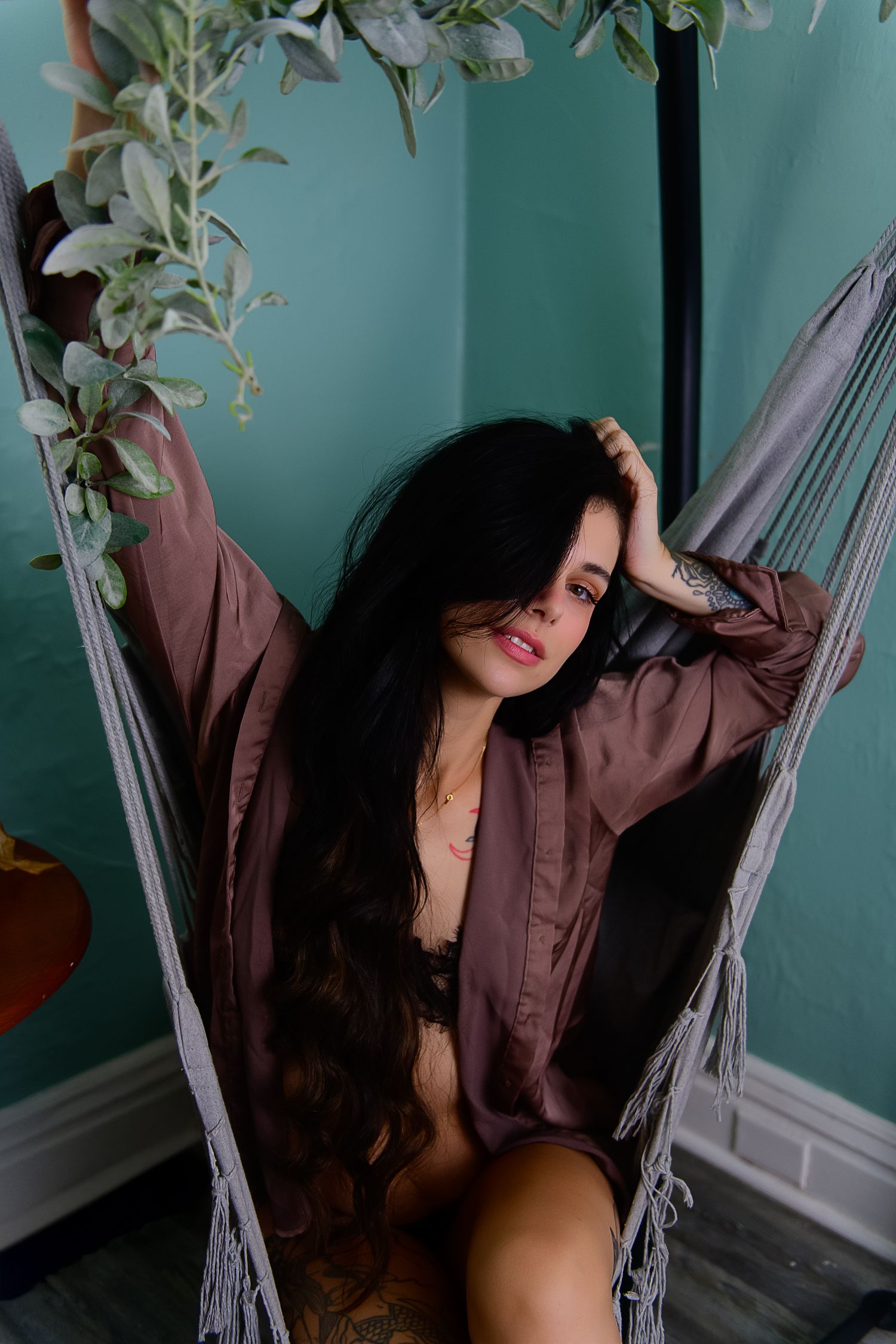 A person relaxes in a hammock chair against a turquoise wall with hanging plants in a boho-style indoor setting.