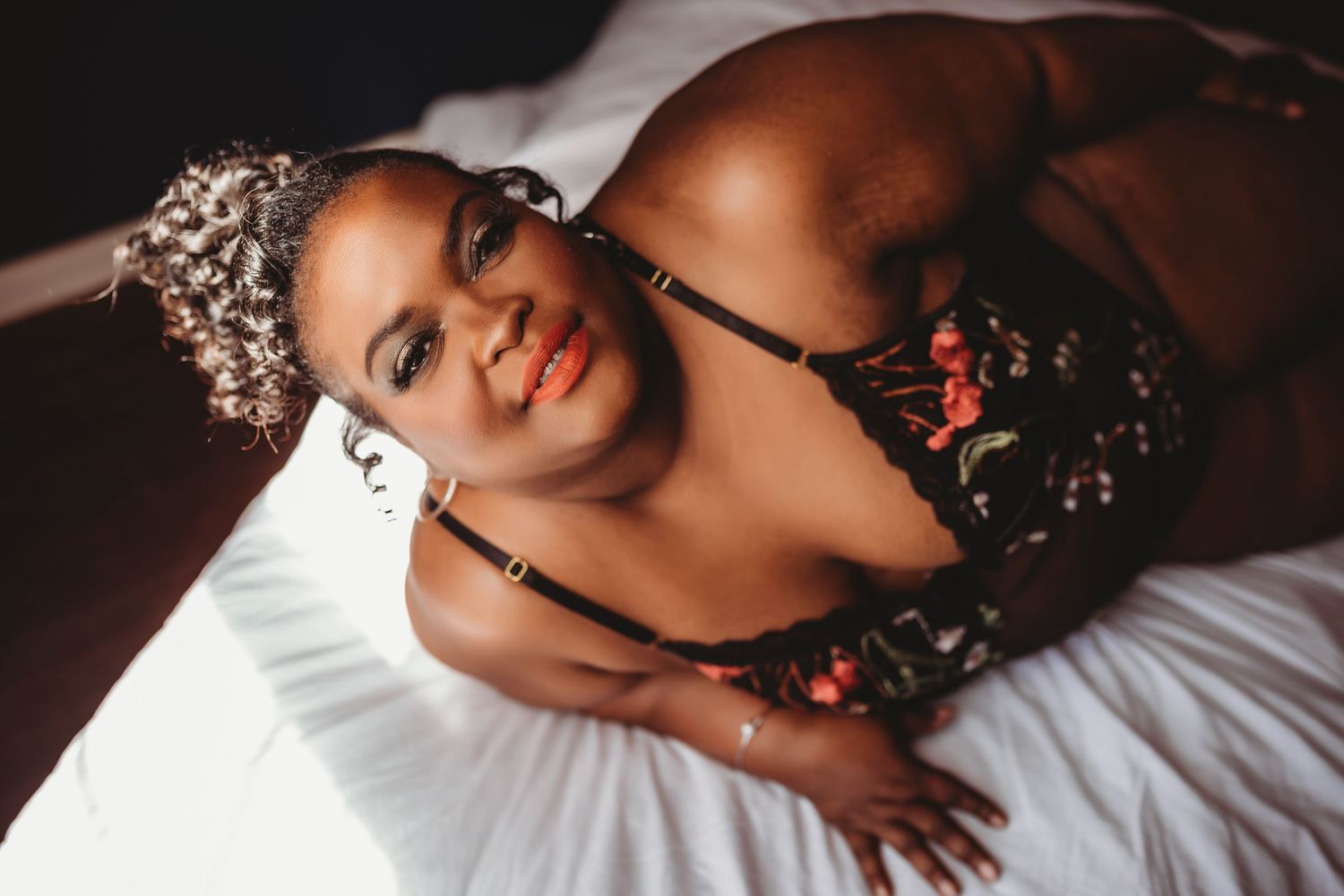A stunning boudoir portrait shows someone wearing a floral lingerie set and sparkly tiara while posing on white bedding.