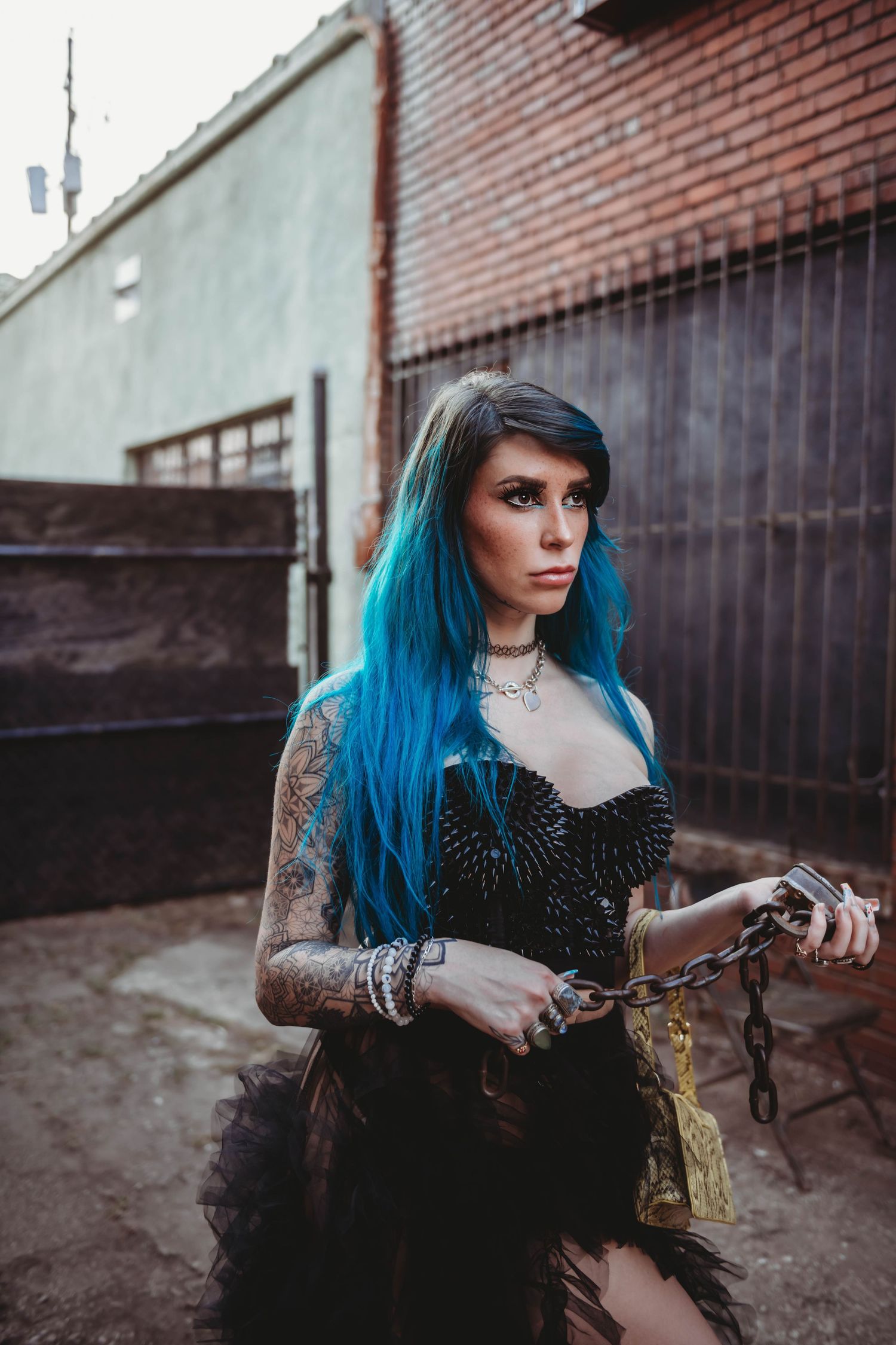 Alternative model with blue hair wearing studded black outfit poses by brick wall.