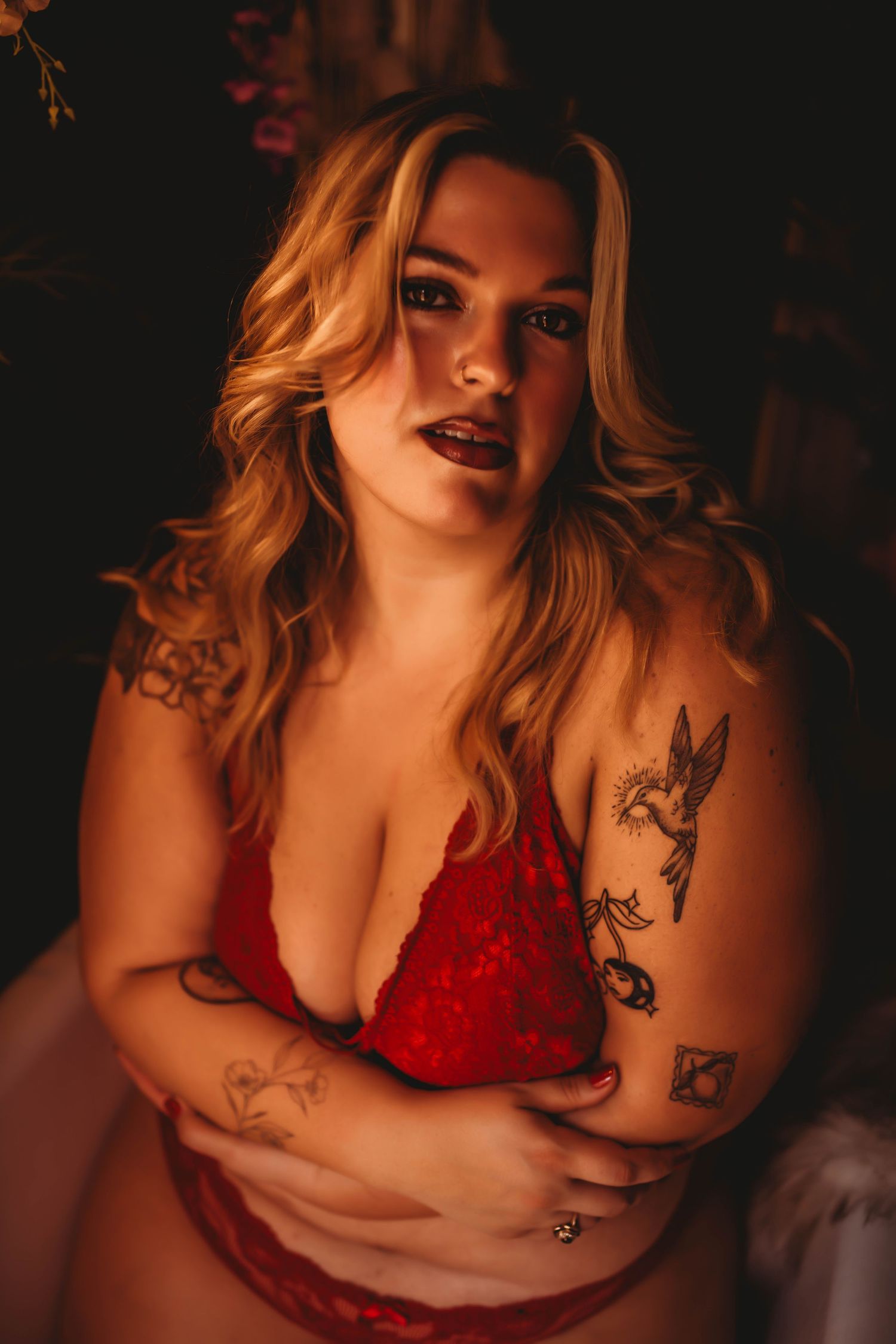 Woman in red lace lingerie with tattoos posing in warm moody lighting for a boudoir photography session.