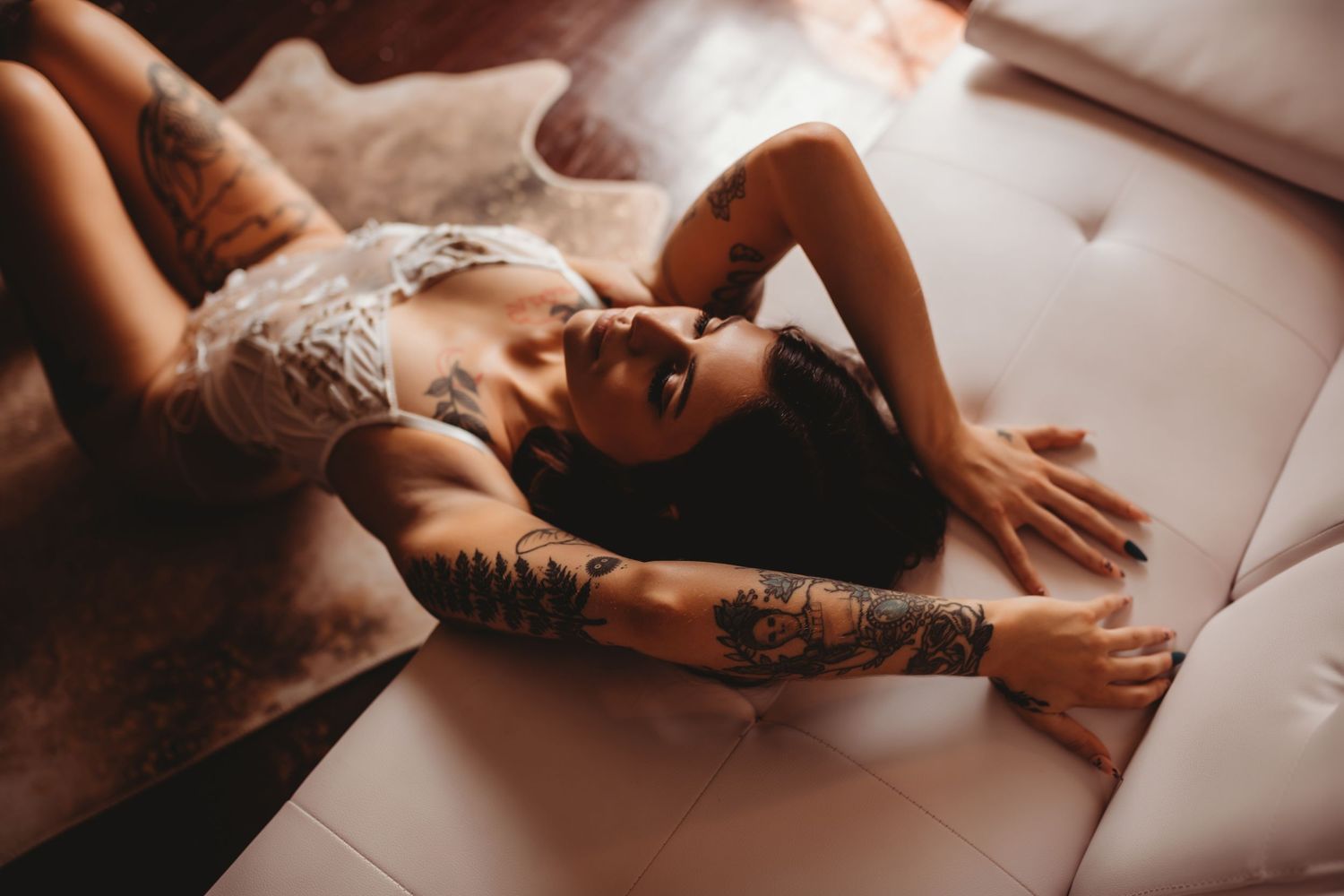 A person in white lace lingerie with visible tattoos reclines seductively on a light colored couch in warm lighting.