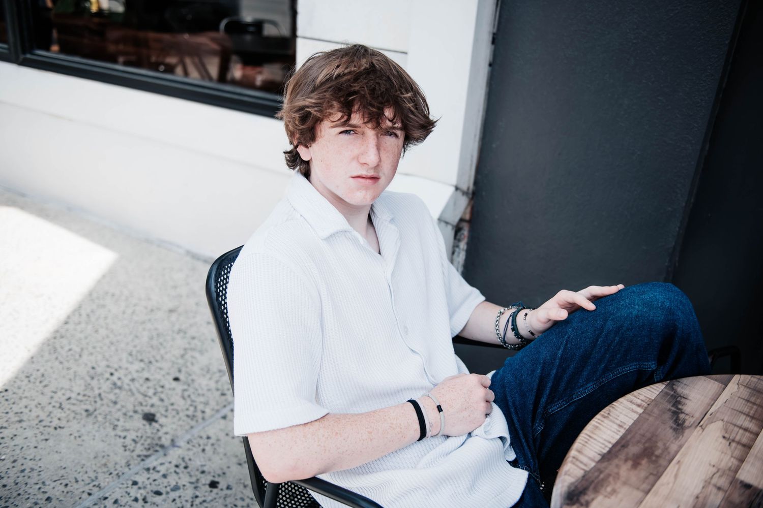 A person in a white button-down shirt and blue jeans sits casually against a wall in natural lighting.