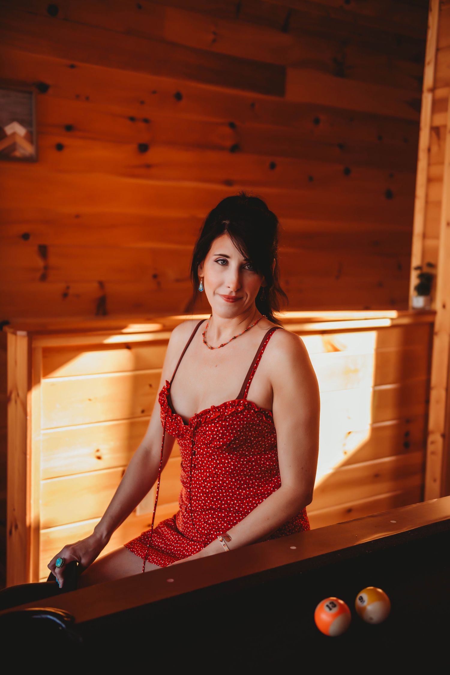 Woman in red polka dot dress poses at pool table in warm wooden cabin setting.