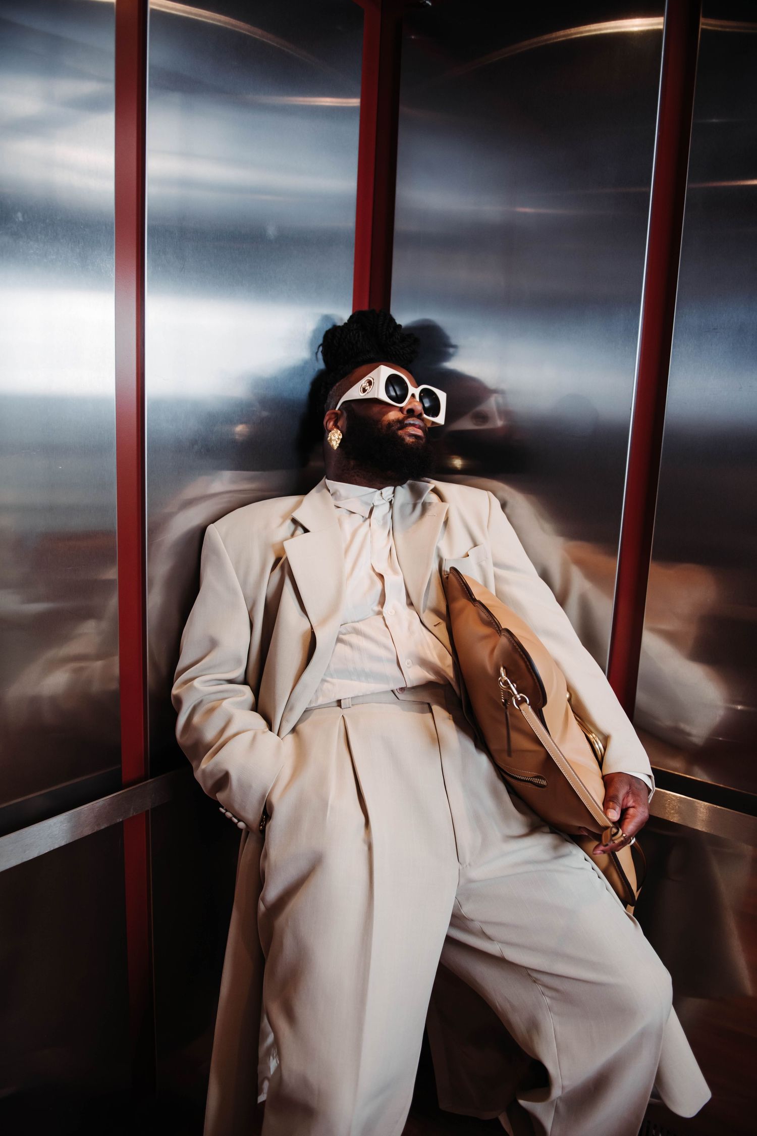 Person in cream colored suit and sunglasses lounging in metallic elevator interior.