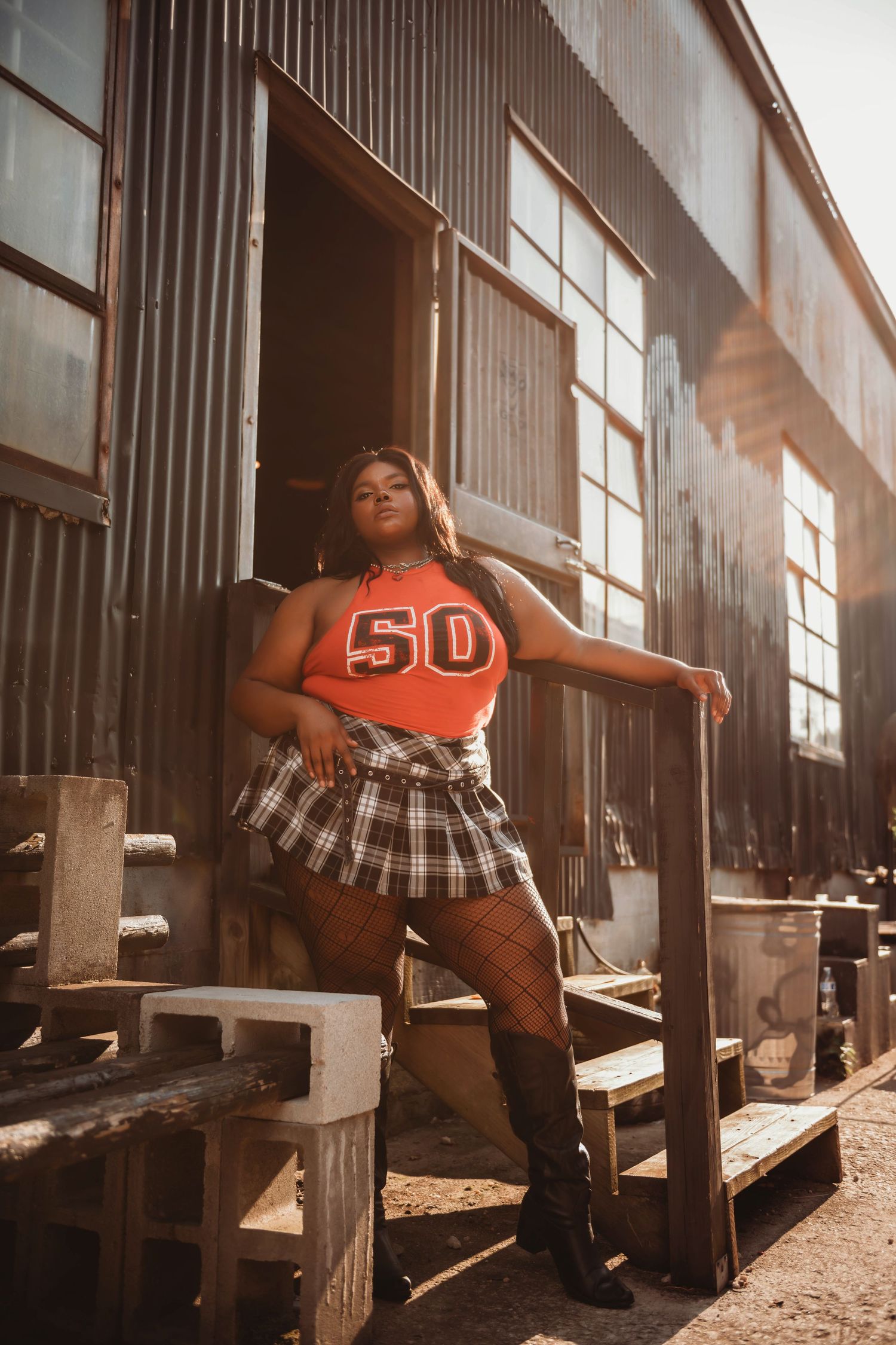 Fashion portrait with orange sports jersey and plaid skirt against industrial backdrop.