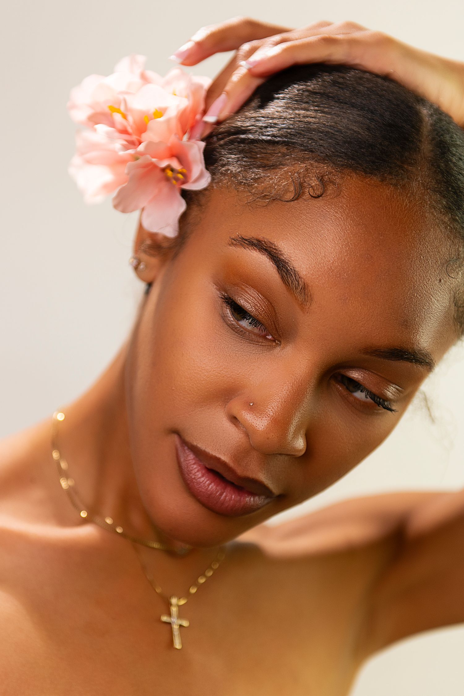 A close-up beauty portrait with soft lighting shows delicate pink flowers and a gold cross necklace against glowing skin.