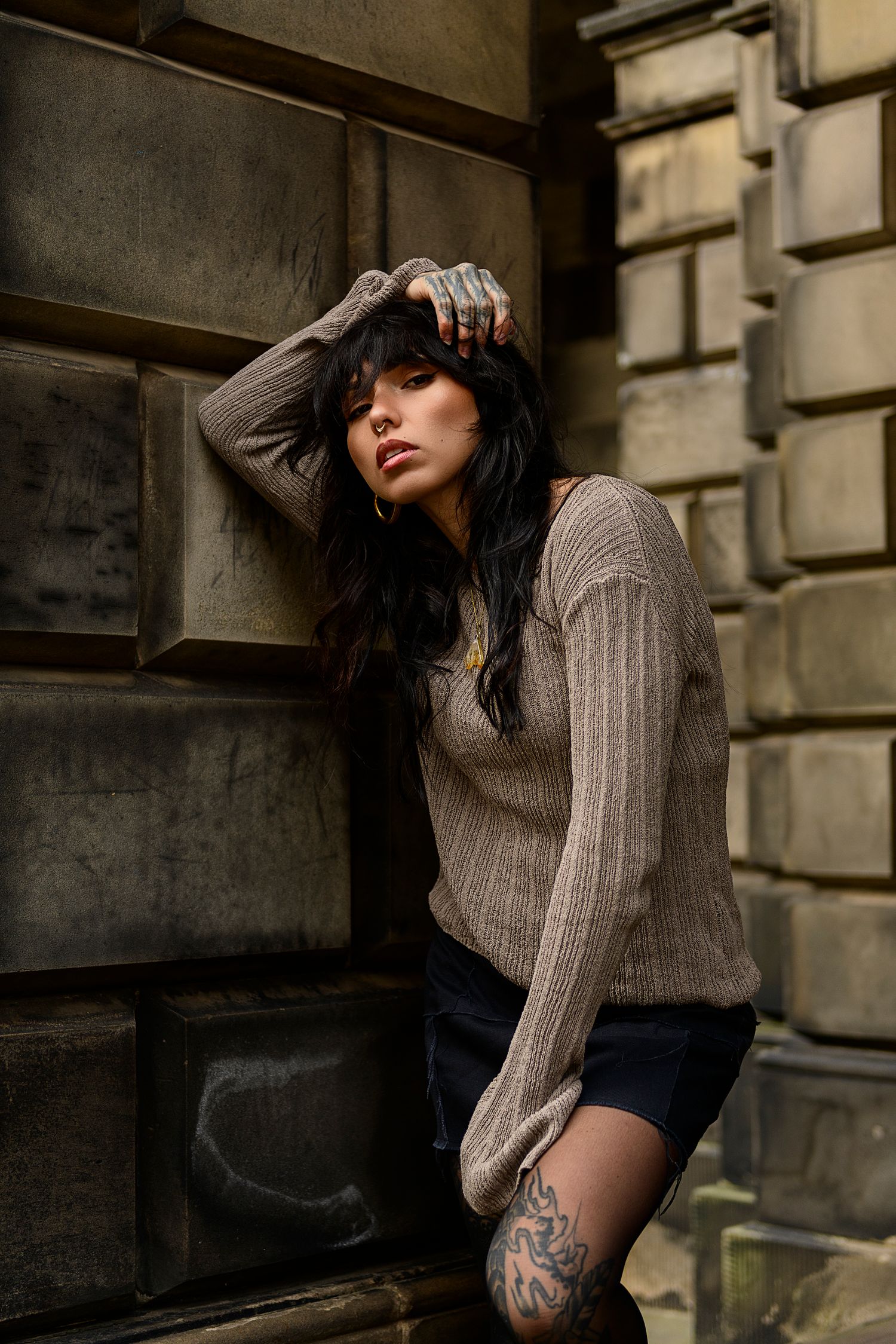 A person in a beige sweater sits against a stone wall in a moody, artistic fashion photoshoot.