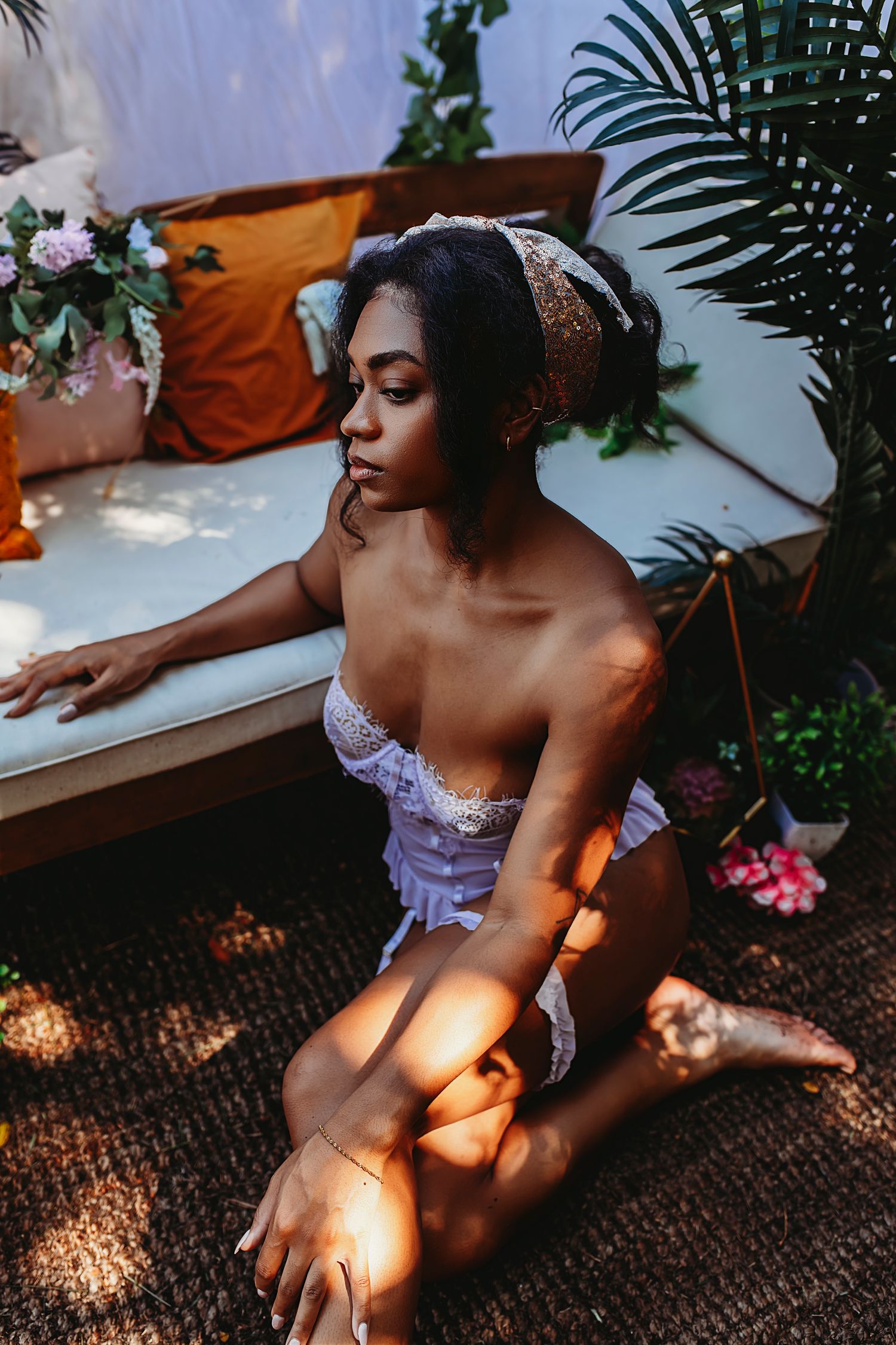 A person in a lavender lace dress sits on a rug in dappled sunlight with tropical plants and decor in the background.