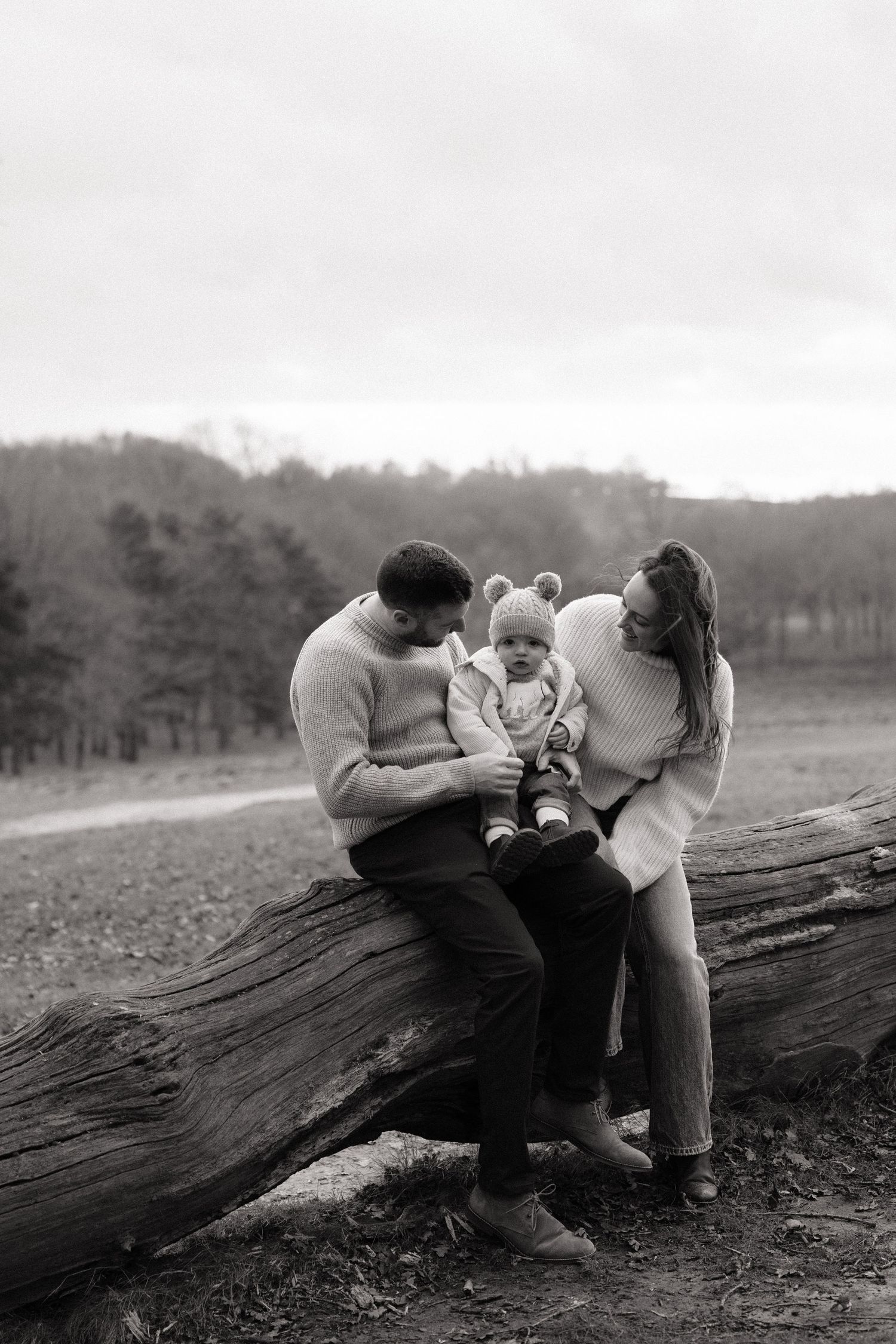 black and white photo of a family of 3 sitting on a fallen down tree trunk cuddled up together