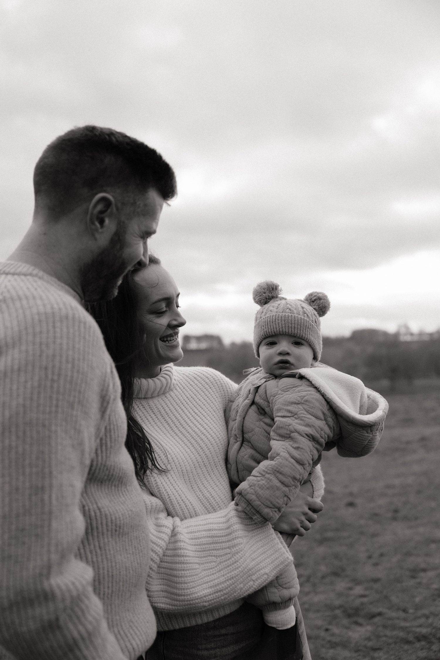 Black and white photo of a smiling mother and father looking at their 15 month old son.