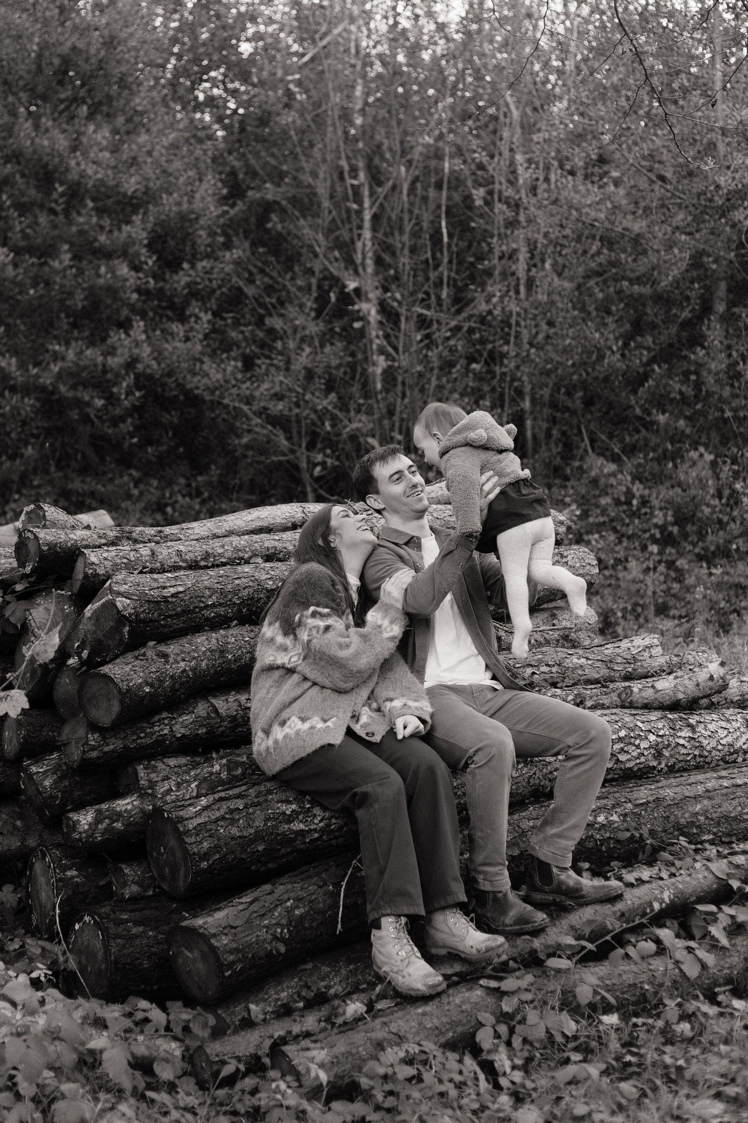 black and white photo of a mother and father sitting on stacked logs holding their baby daughter up so she faces them and smiling at her