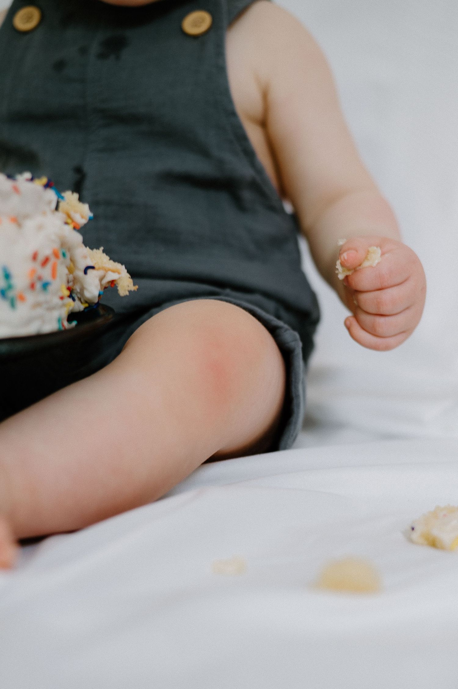 Small child in dark overalls sitting on white bedding with cake crumbs.