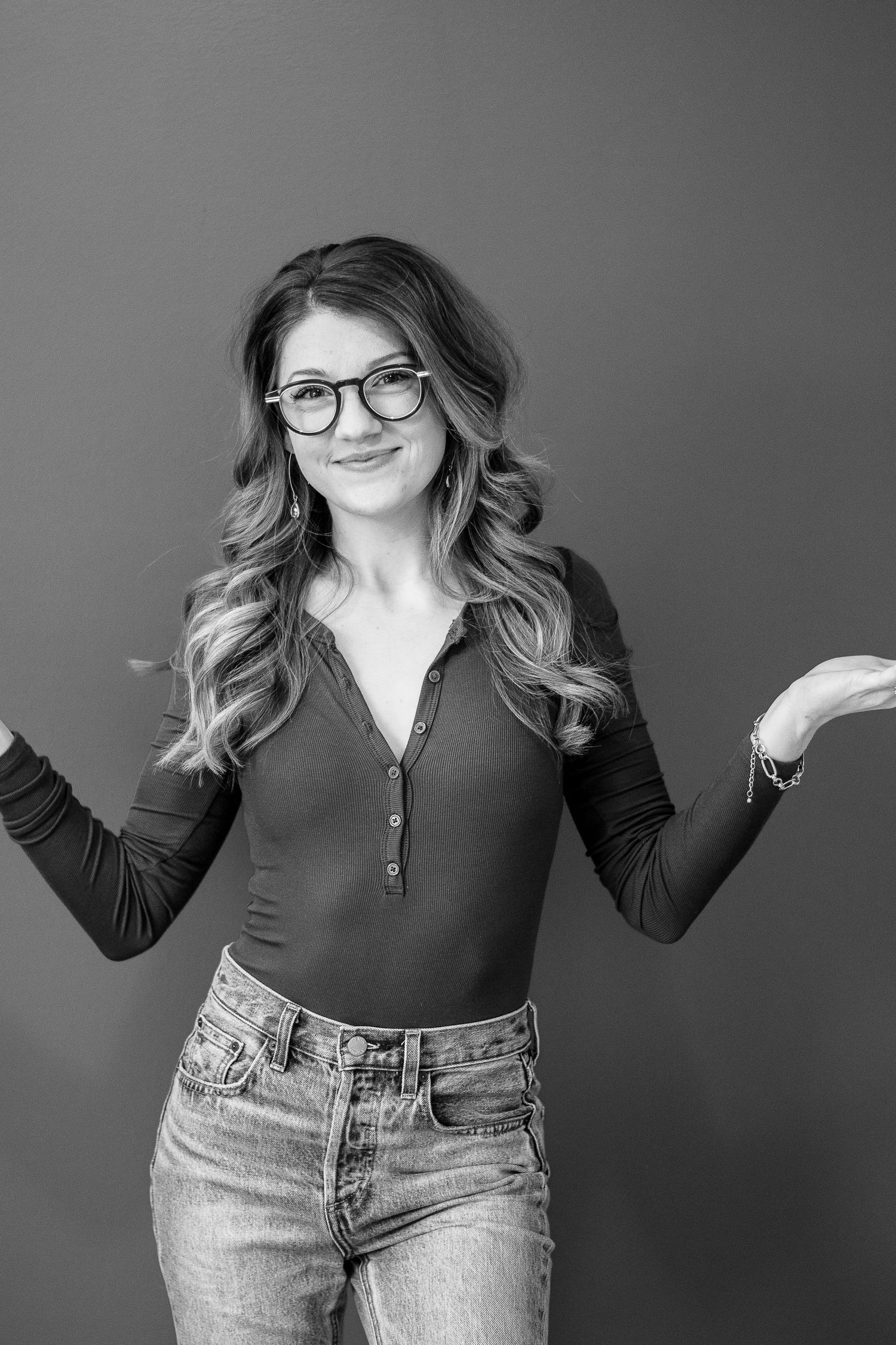Woman in glasses and dark top poses playfully against grey background in studio portraits.