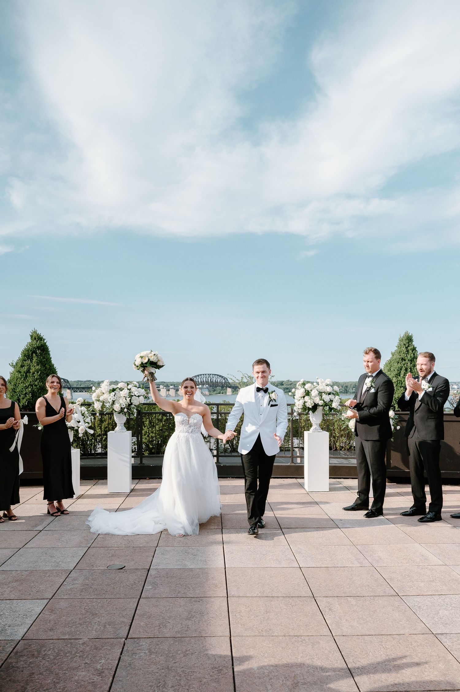 Bride and groom walk down the aisle at an elegant rooftop wedding ceremony with white floral arrangements.