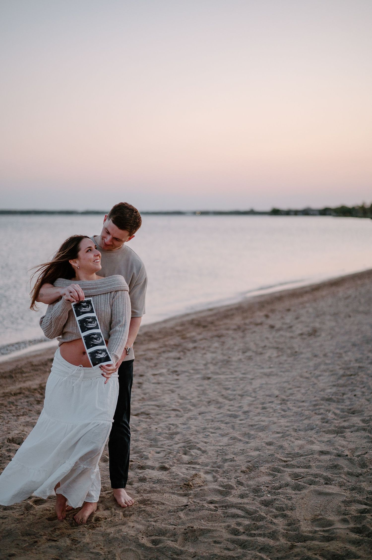 Couple shares pregnancy news with ultrasound photos during sunset beach photoshoot.