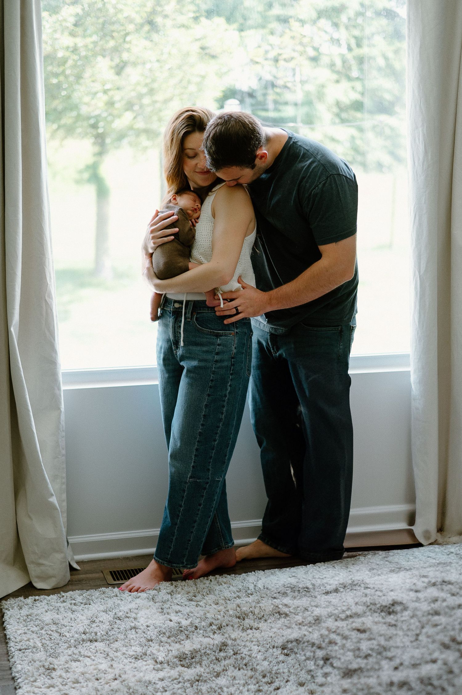 A couple shares an intimate moment while holding their baby by a window.