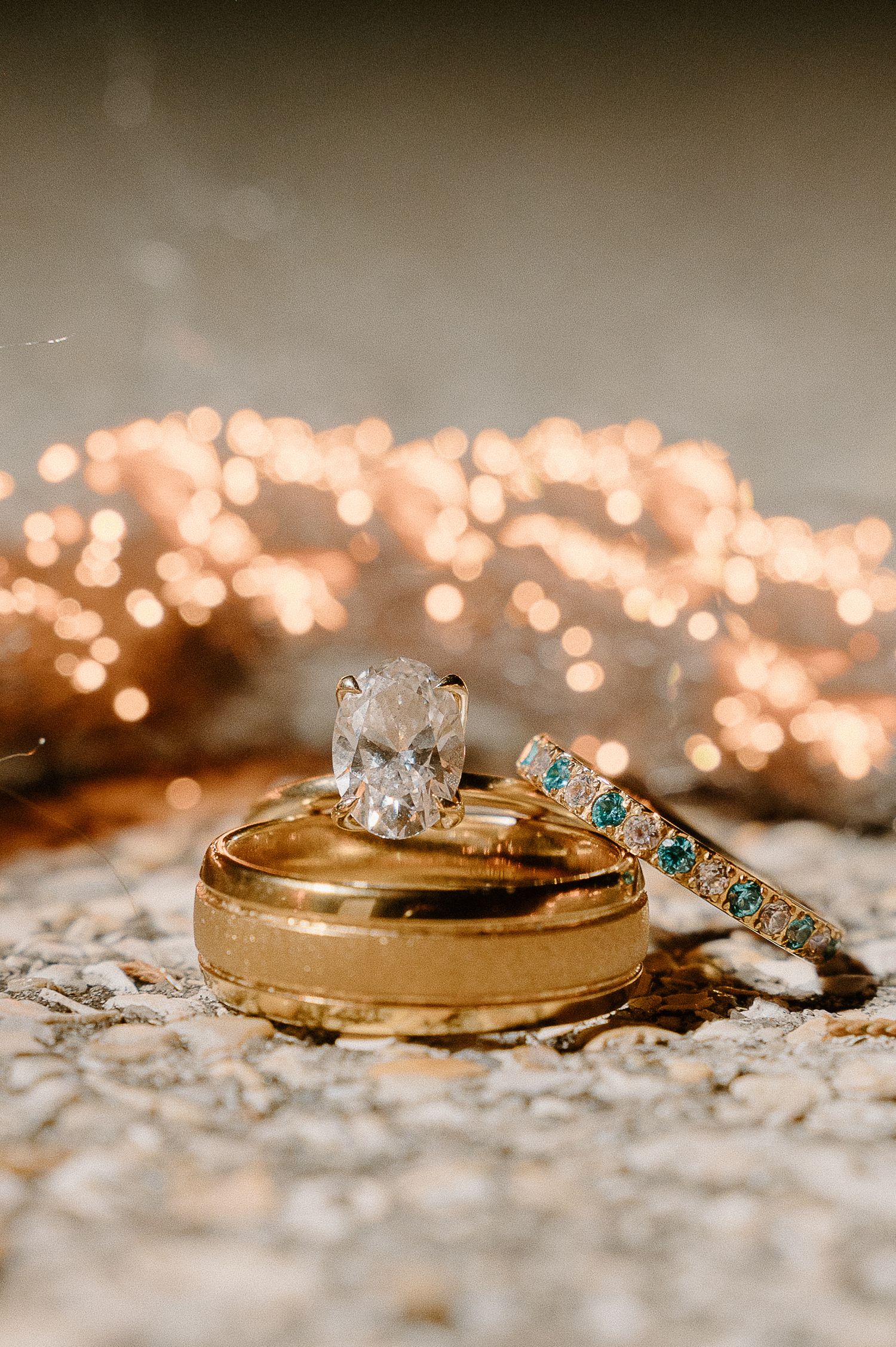 Gold wedding band, diamond oval engagement ring, and teal gemstone eternity band stacked together with bokeh lights background.