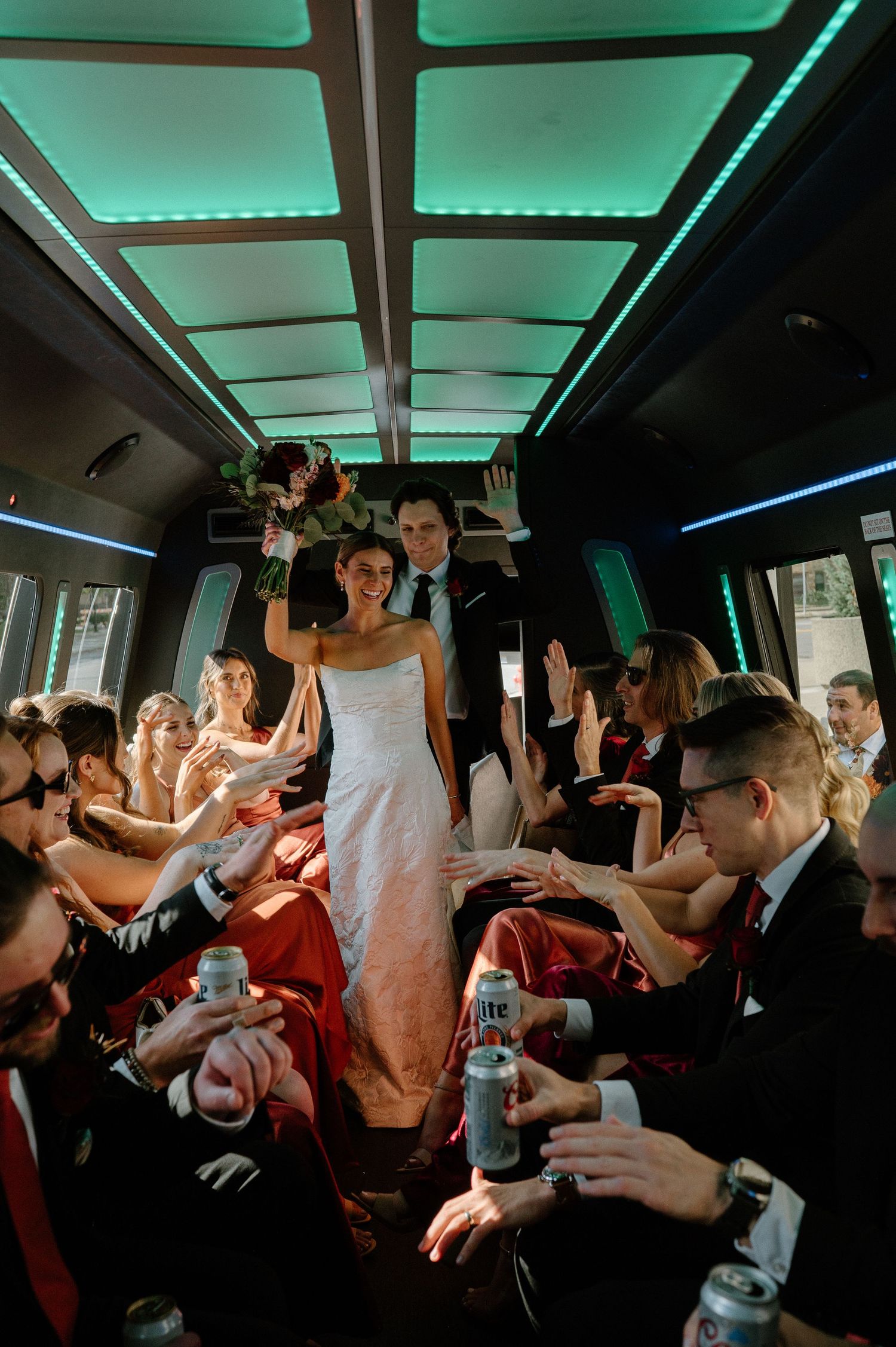 Wedding party guests dance and celebrate inside a party bus illuminated with turquoise LED lighting.