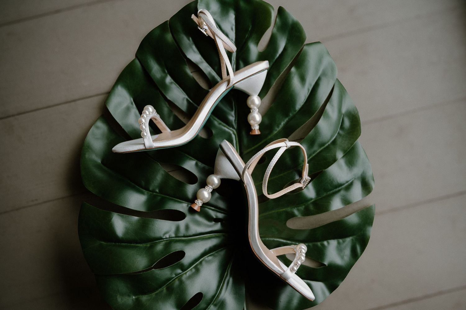 Elegant white pearl-embellished strappy heeled sandals displayed on a tropical monstera leaf on wooden floor.