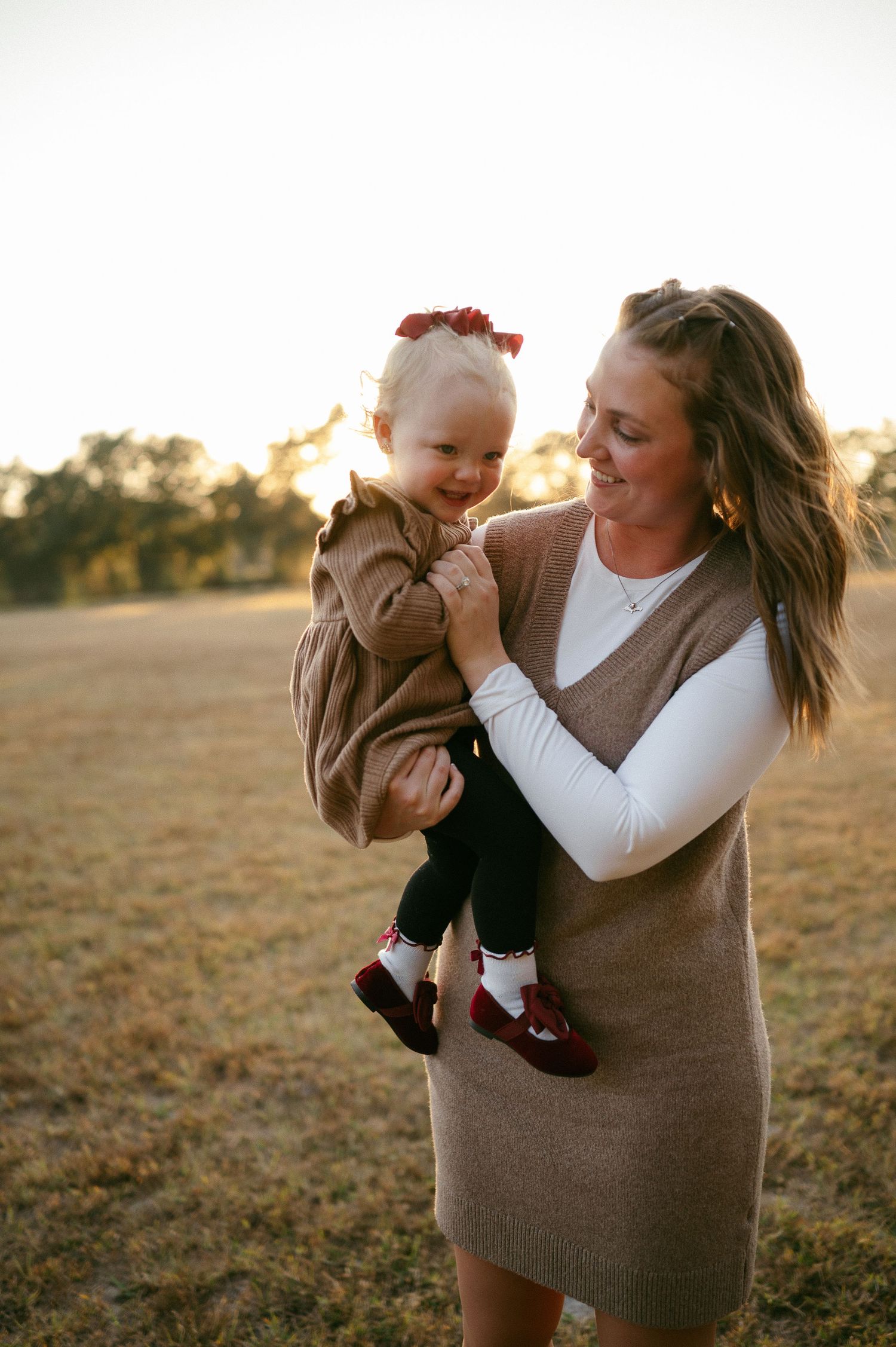 A smiling woman holds a happy toddler girl in a brown sweater outdoors during golden hour.