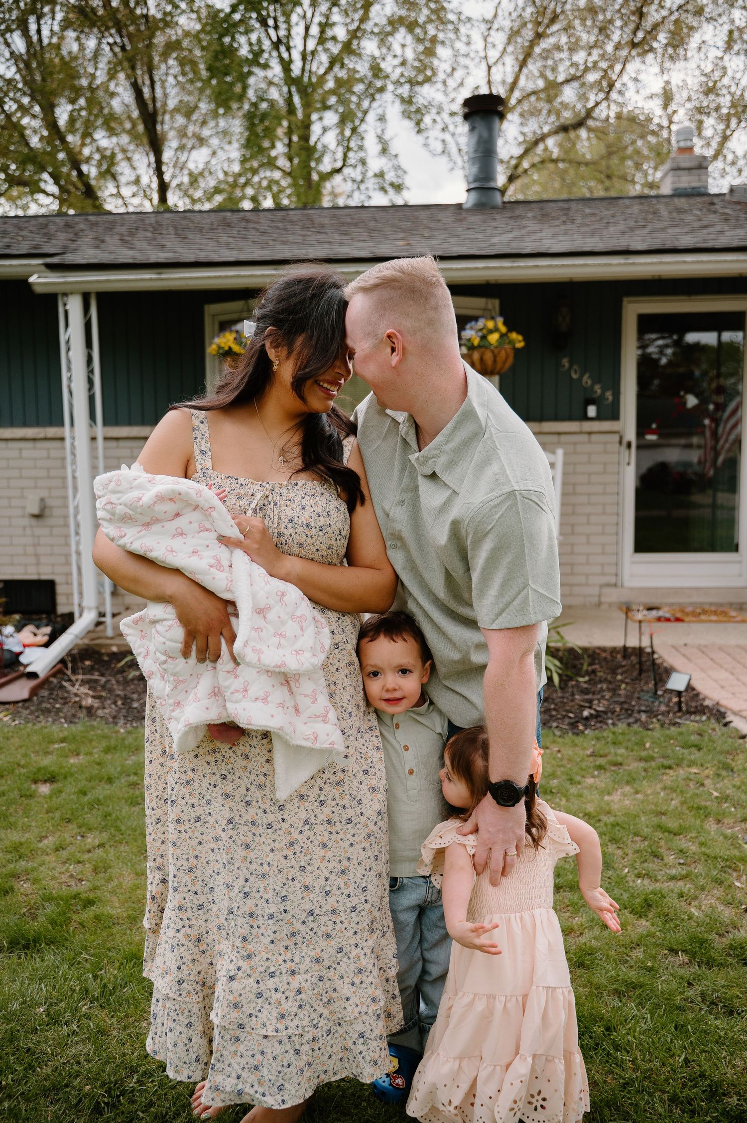 Young family of four with newborn baby poses lovingly in front of cozy home, surrounded by lush green lawn.
