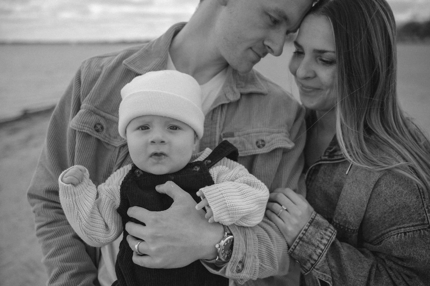 Black and white photo of a young couple holding their baby outdoors near a beach, sharing a tender family moment.
