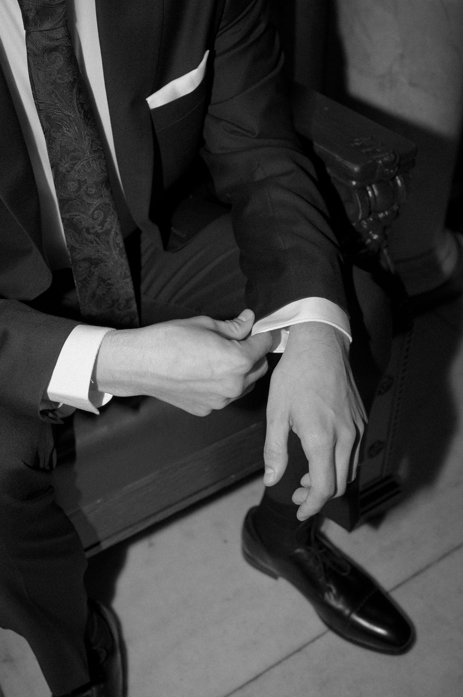 A black and white photo shows hands adjusting formal cuff links while wearing a dark suit and dress shoes.